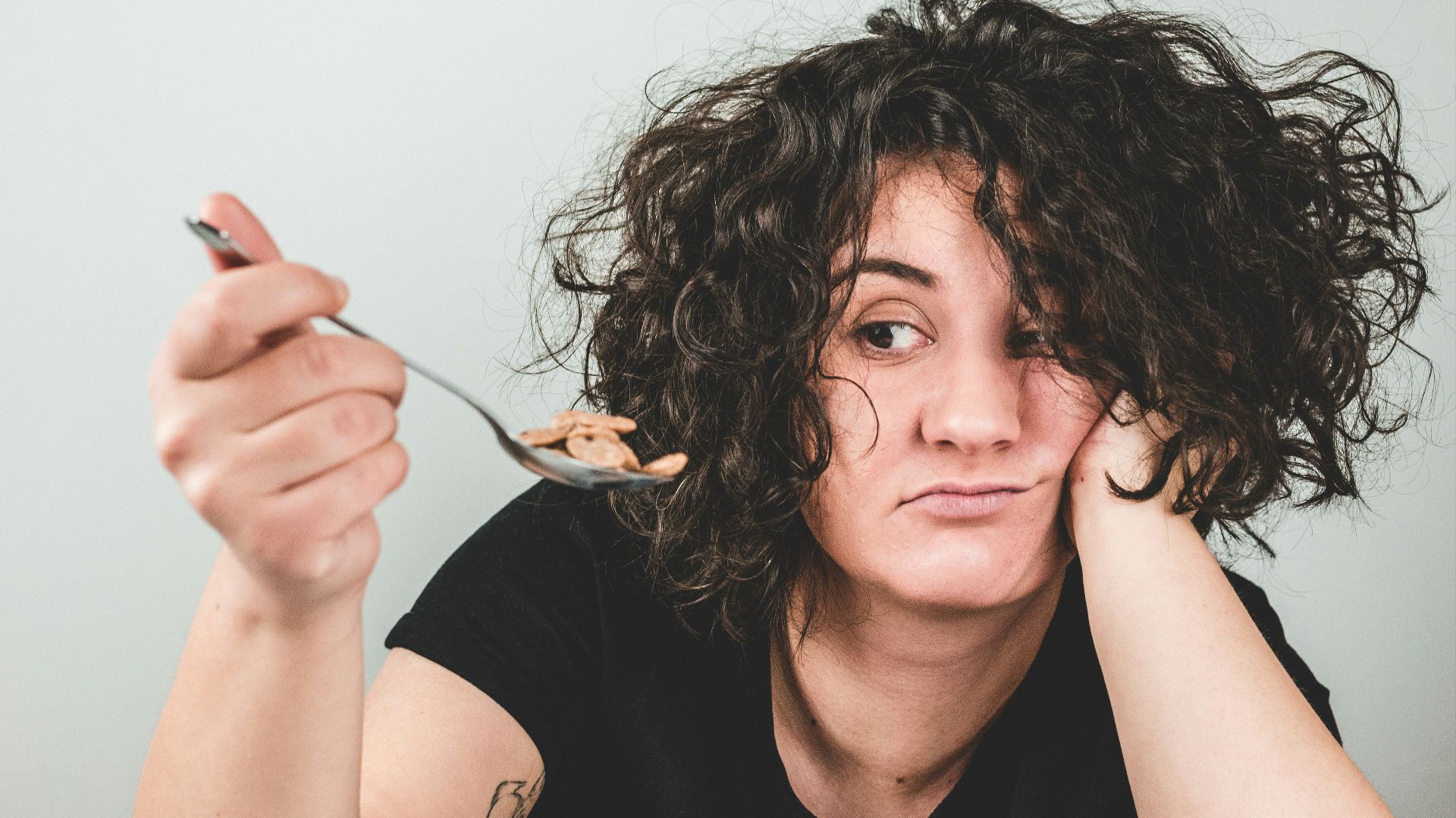 woman with messy hair wearing black crew-neck t-shirt holding spoon with cereals on top