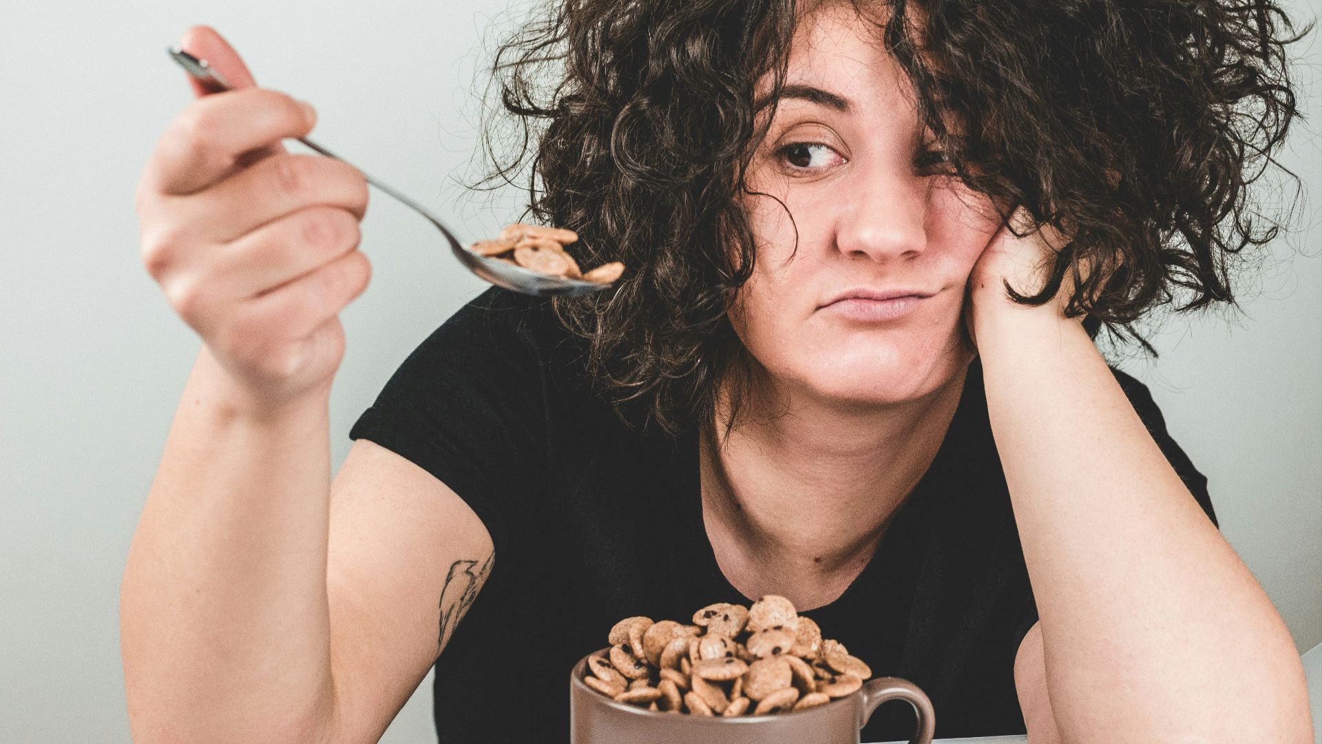 woman with messy hair wearing black crew-neck t-shirt holding spoon with cereals on top