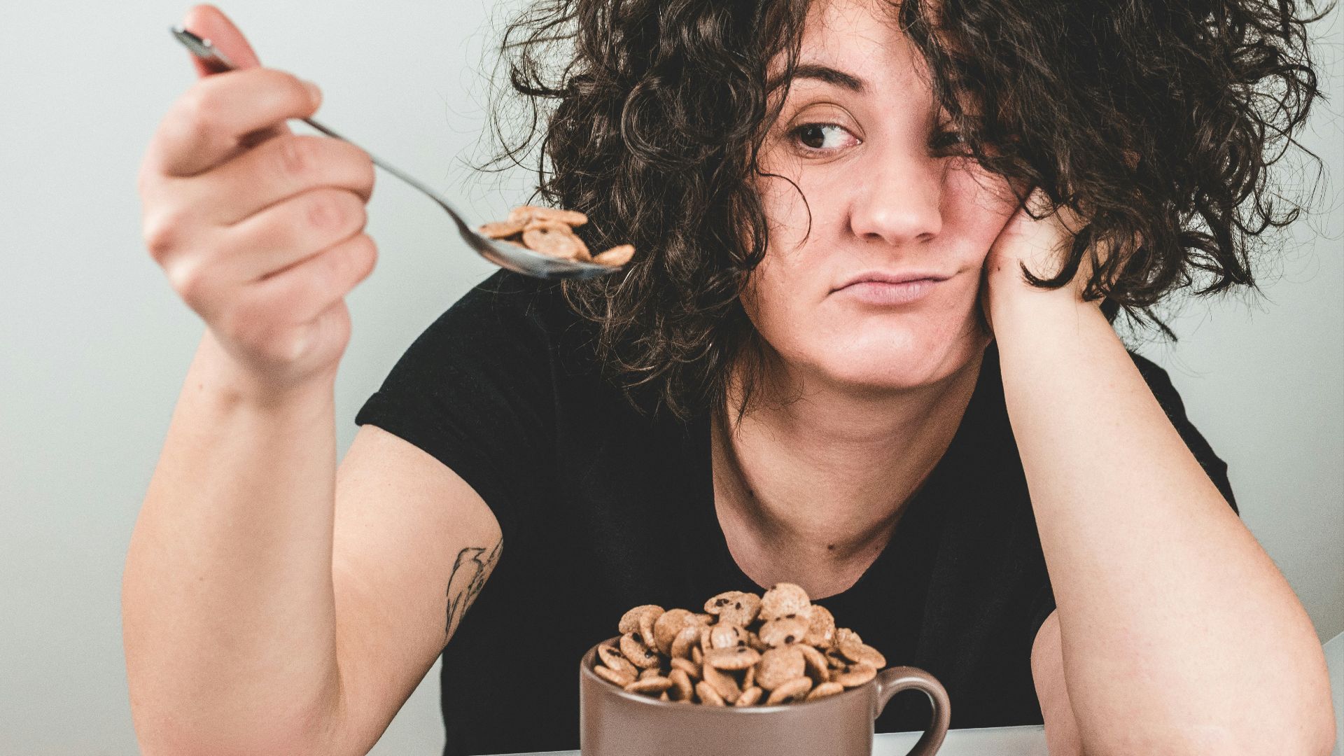 woman with messy hair wearing black crew-neck t-shirt holding spoon with cereals on top