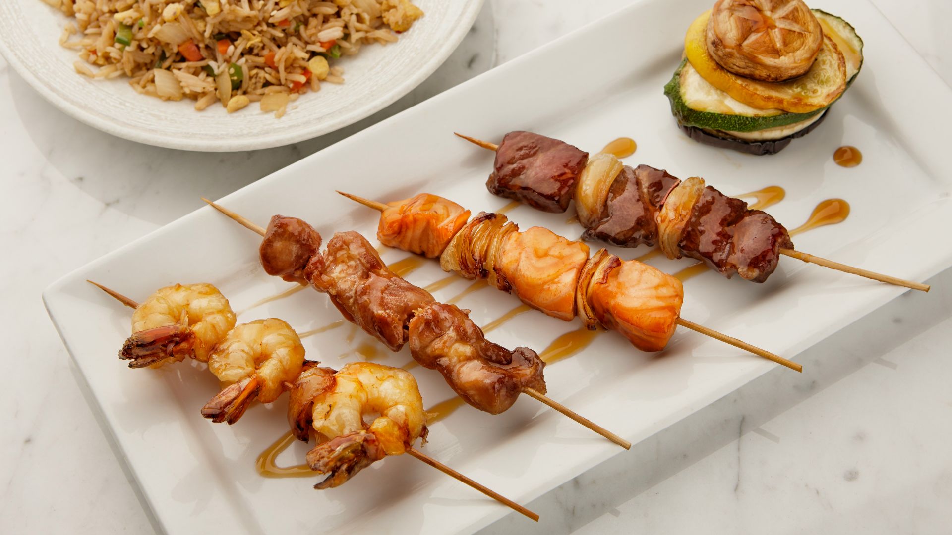 a white plate topped with skewers of food next to a bowl of rice