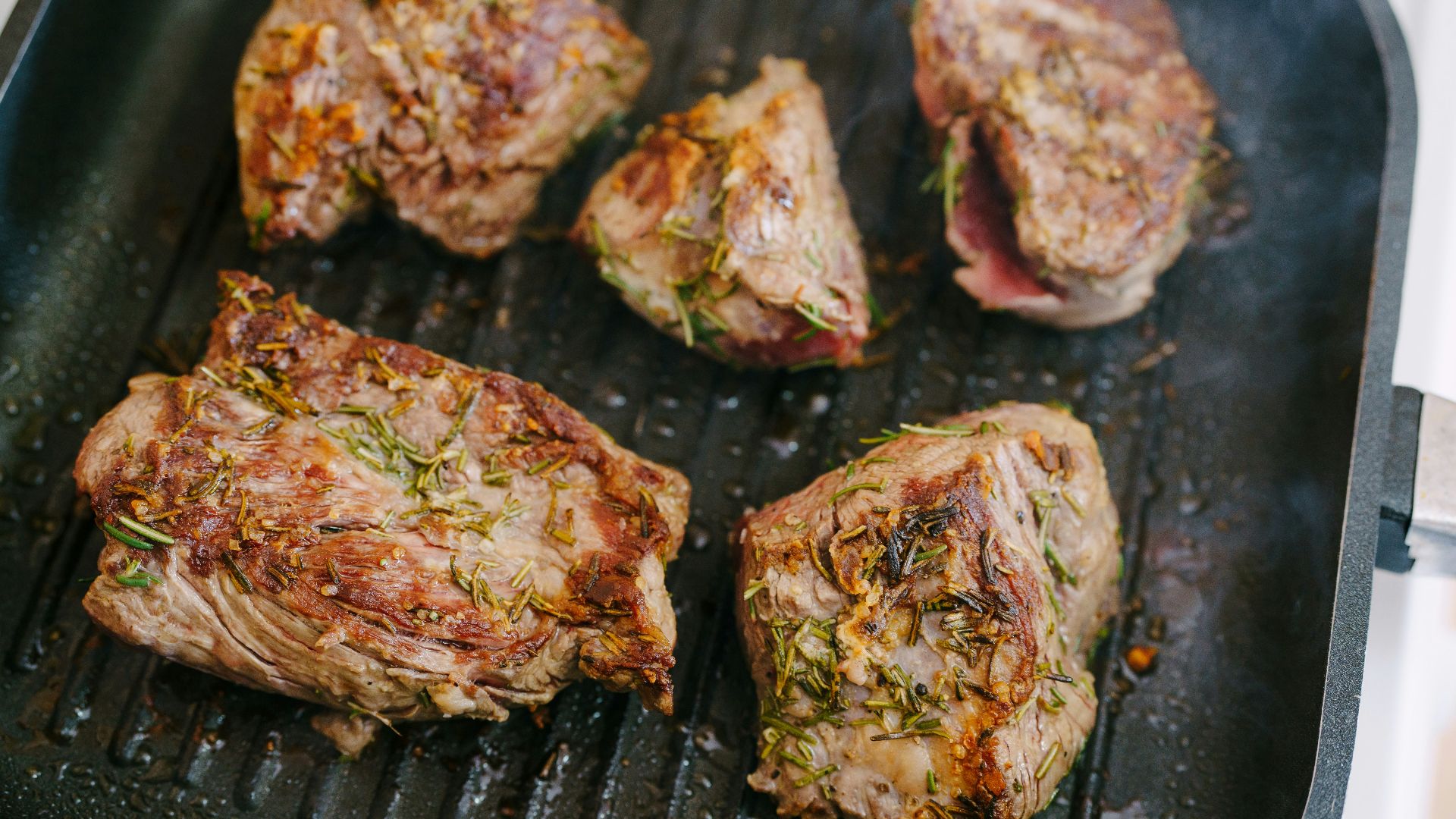 Five pieces of seasoned meat cooking on a grill pan.