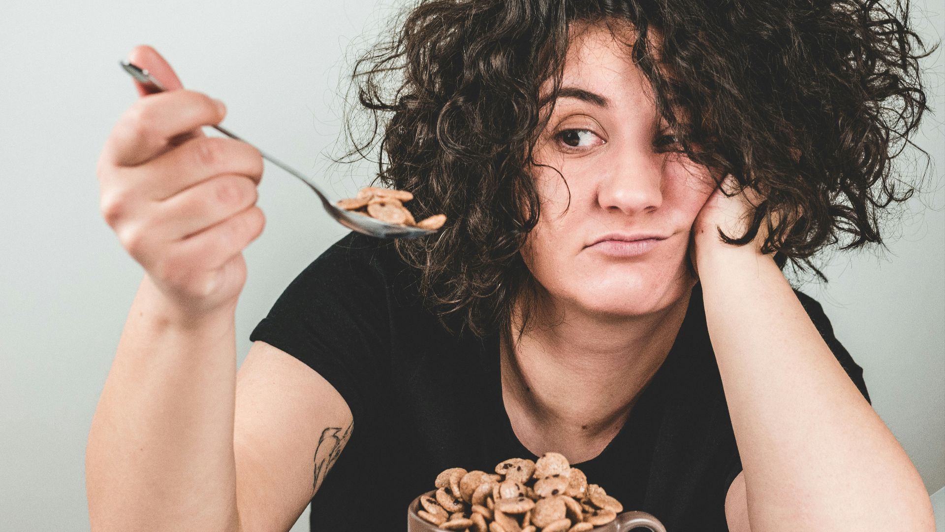 woman with messy hair wearing black crew-neck t-shirt holding spoon with cereals on top