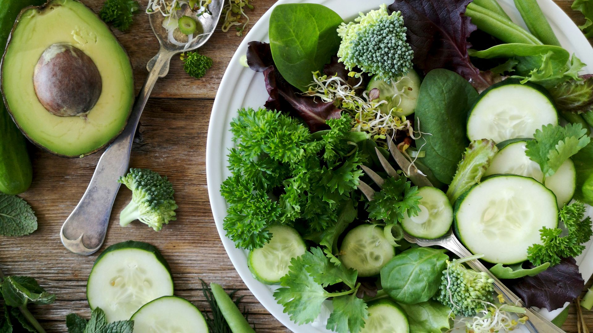 sliced broccoli and cucumber on plate with gray stainless steel fork near green bell pepper, snowpea, and avocado fruit