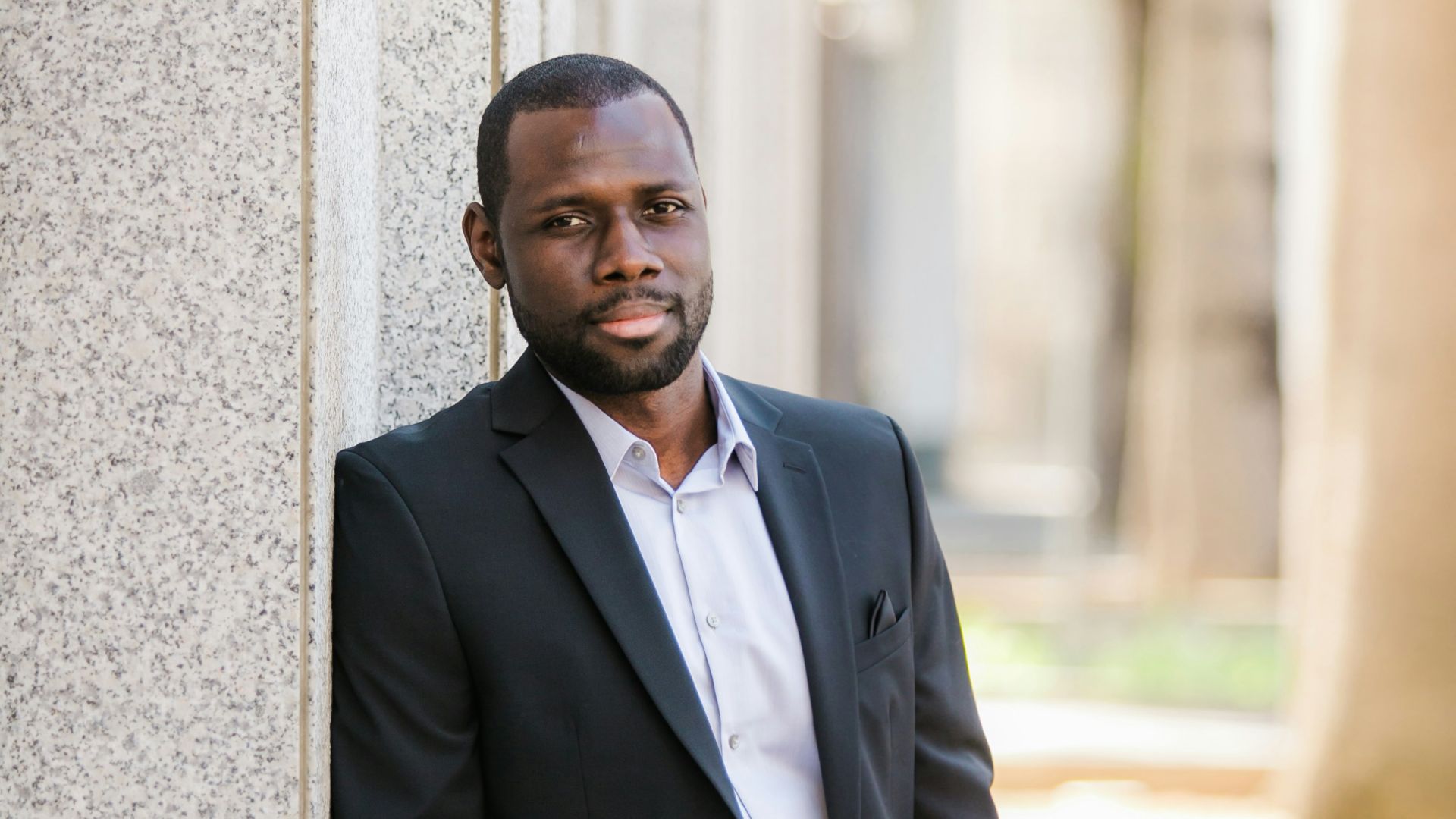 man in black suit jacket leaning on gray concrete wall during daytime