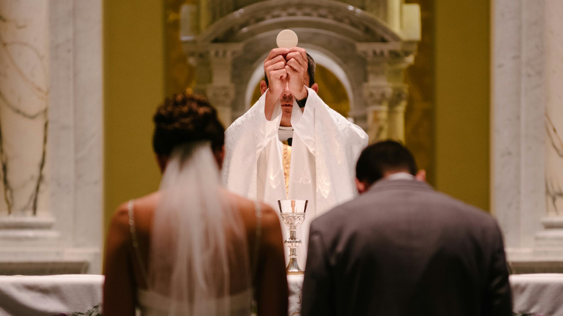 groom and bride kneeling in front of priest raising The Holy Sacrament