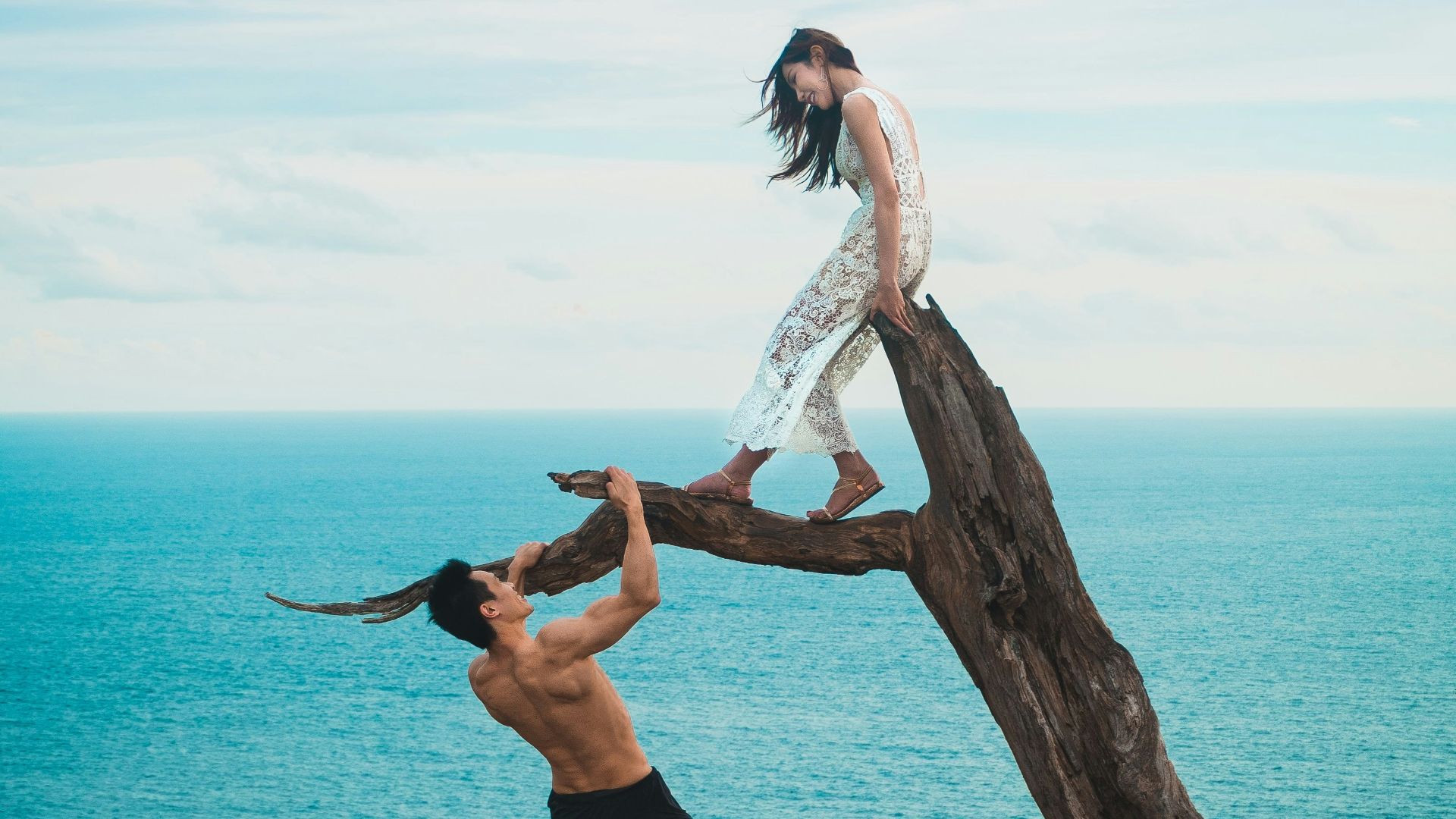woman sitting on tree trunk with man holding on branch near sea under white clouds during daytime