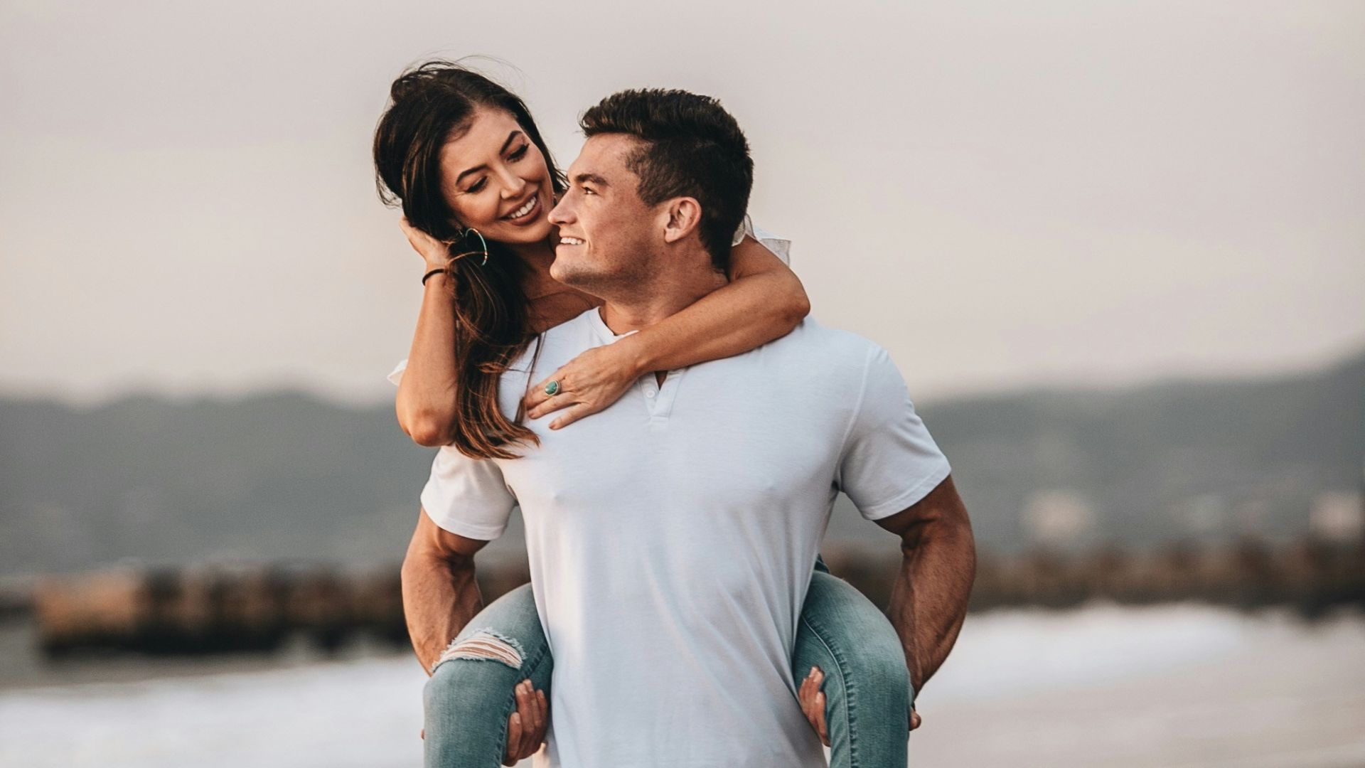 man in white crew neck t-shirt kissing woman in blue denim jeans on beach during