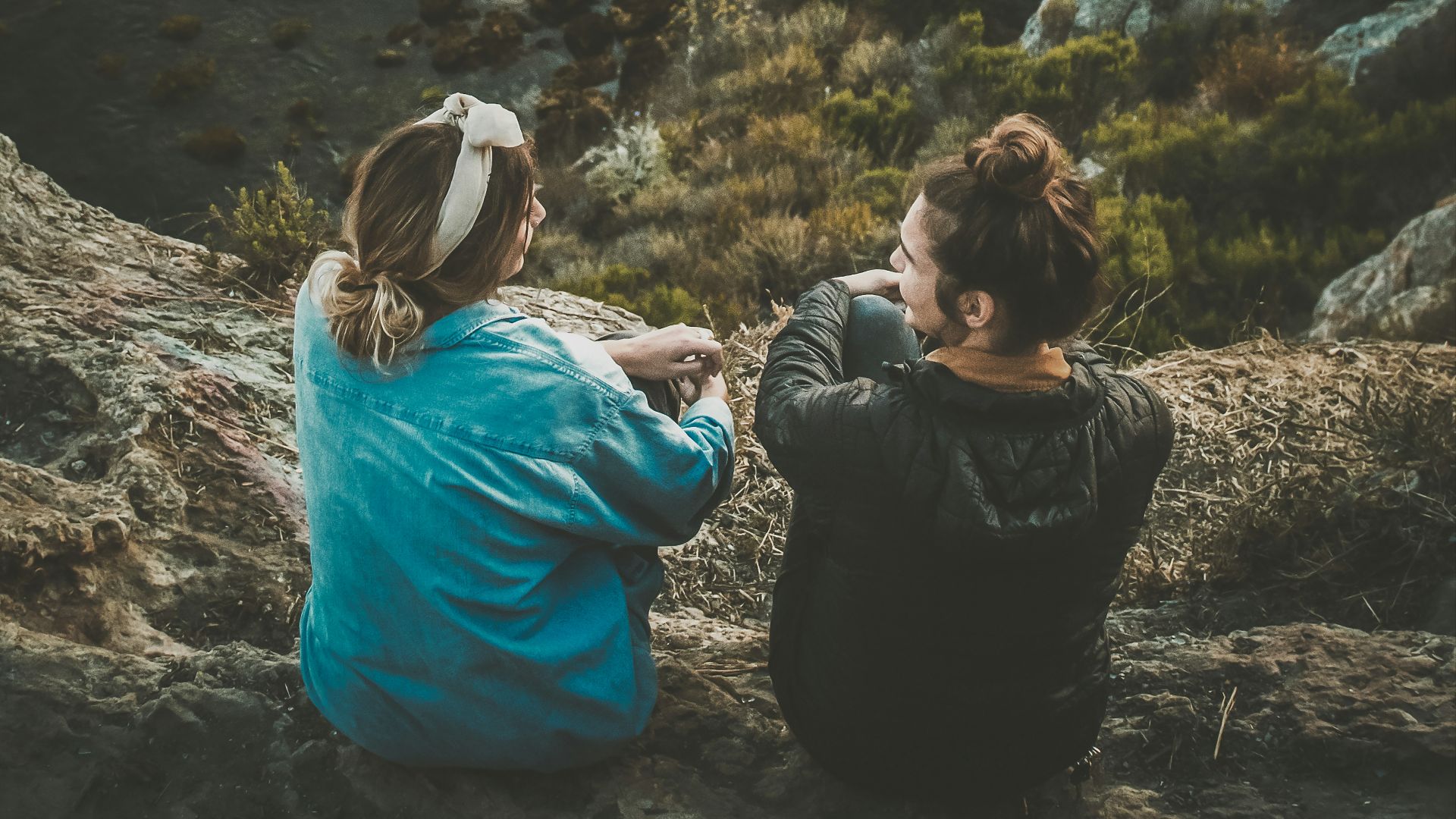 two women sitting on cliff while chatting viewing blue body of water