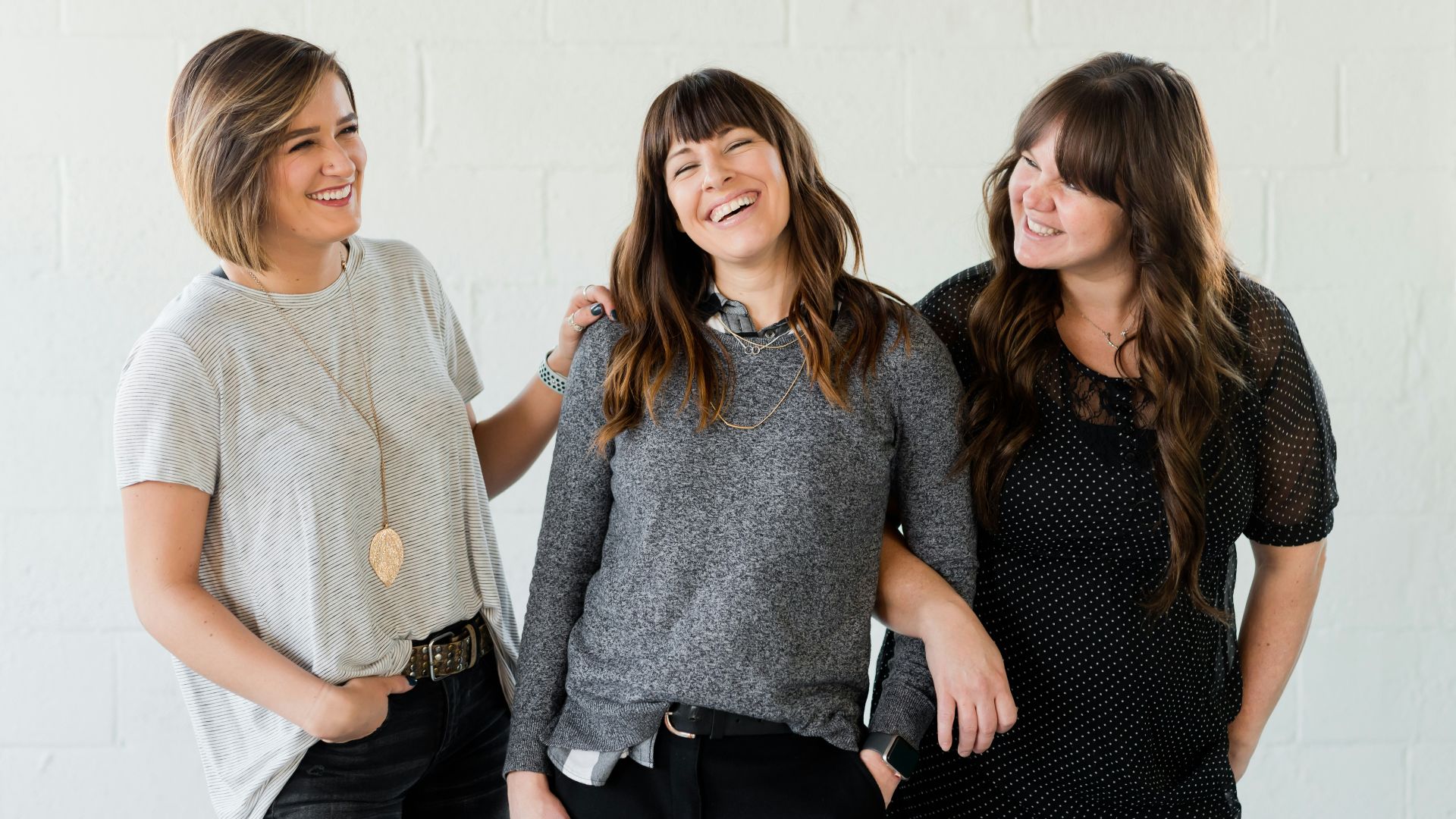 3 women smiling and standing beside white wall