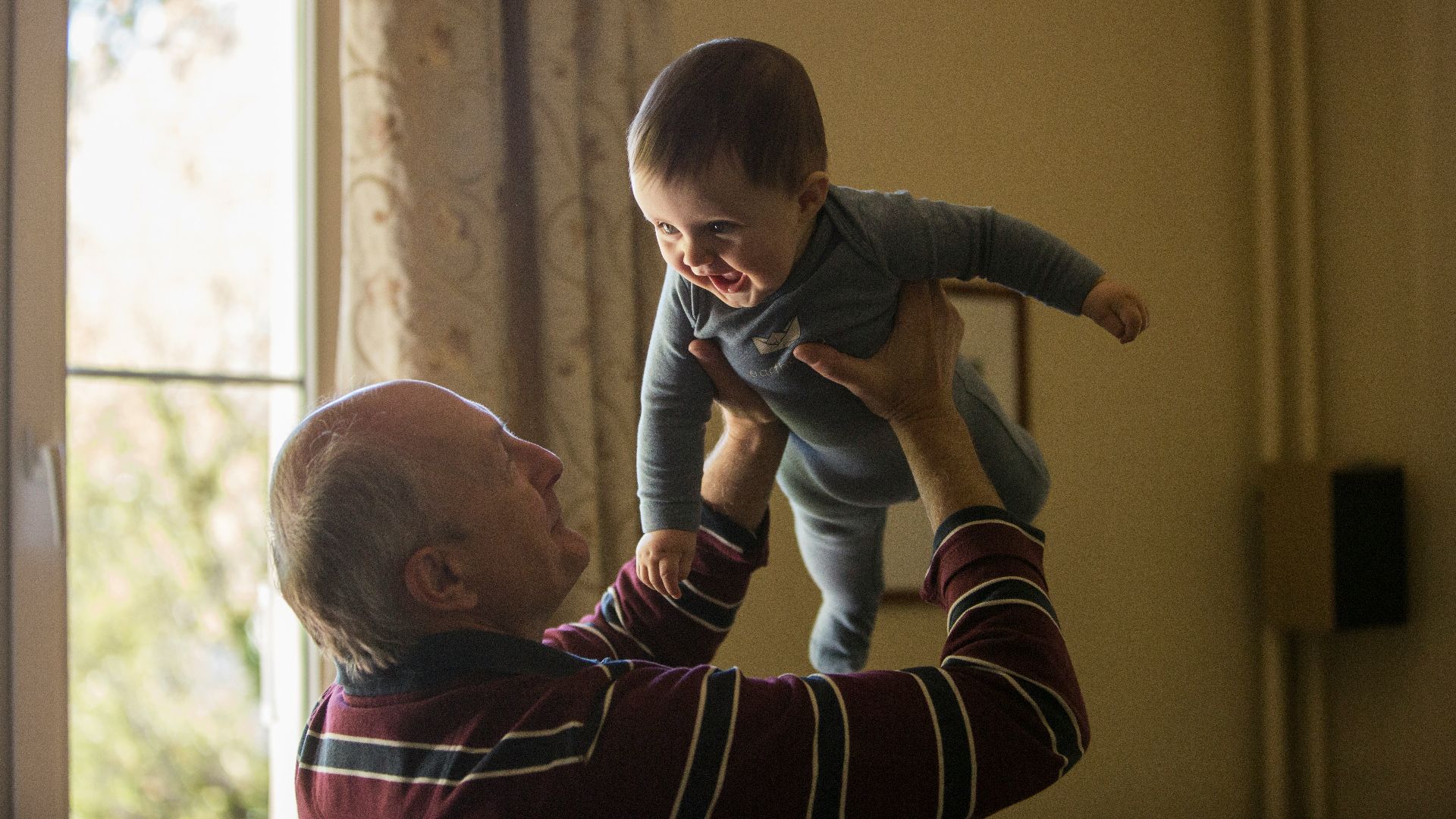 man wearing maroon, white, and blue stripe long-sleeved shirt lifting up baby wearing gray onesie