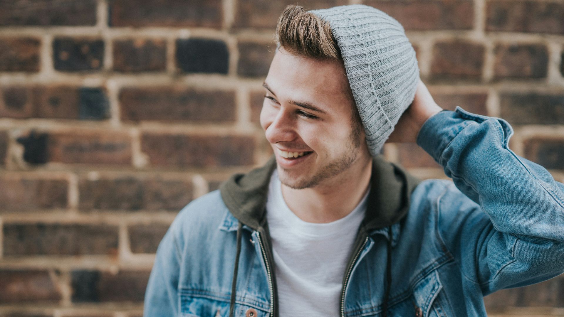 man holding the back of his head while smiling near brick wall