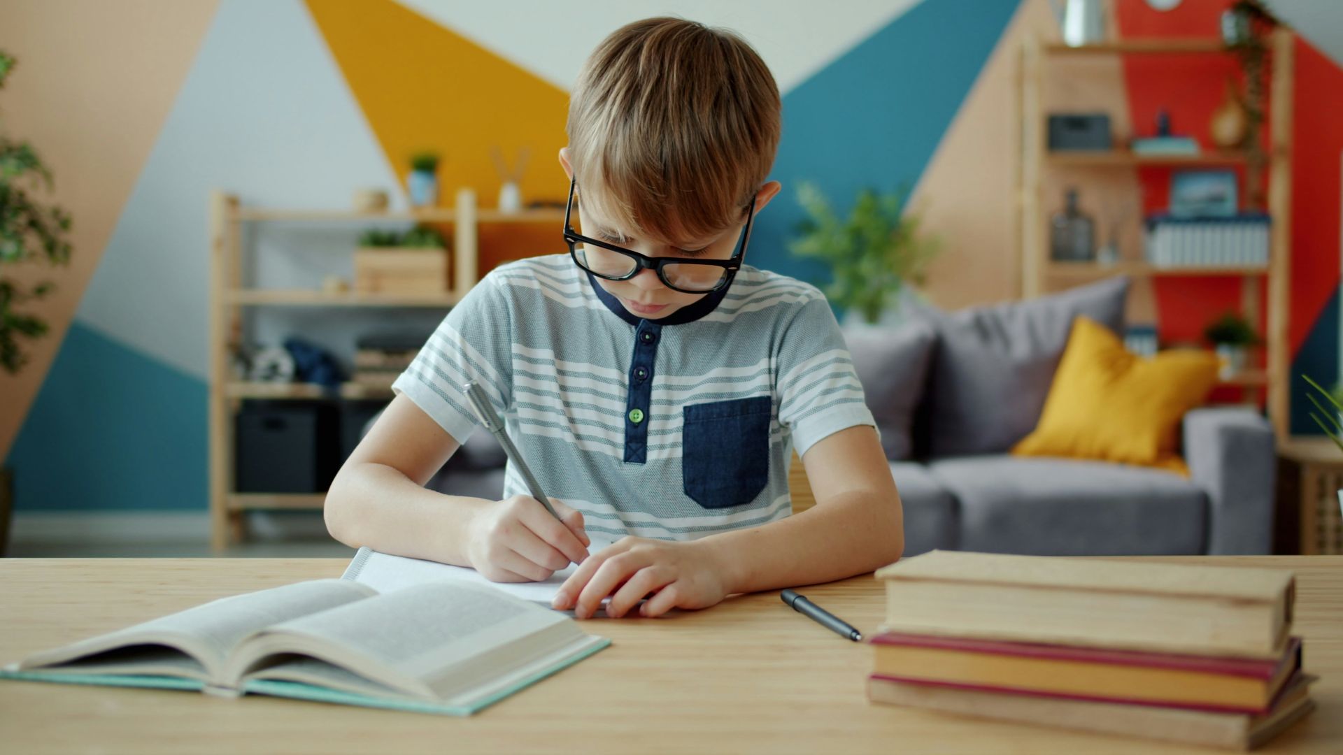 Young boy with glasses writing in a notebook at desk.