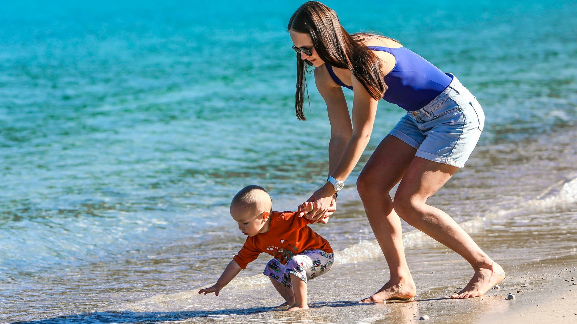 woman in blue and white tank top holding baby in white onesie on beach during daytime
