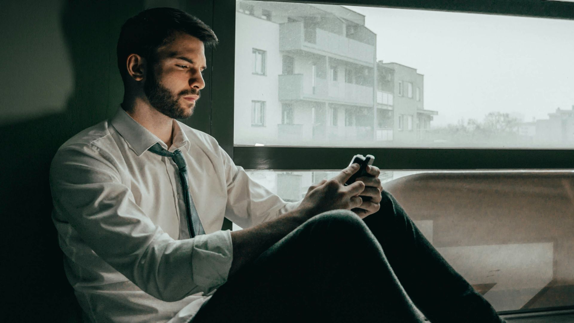 a man sitting on a window sill looking at his cell phone