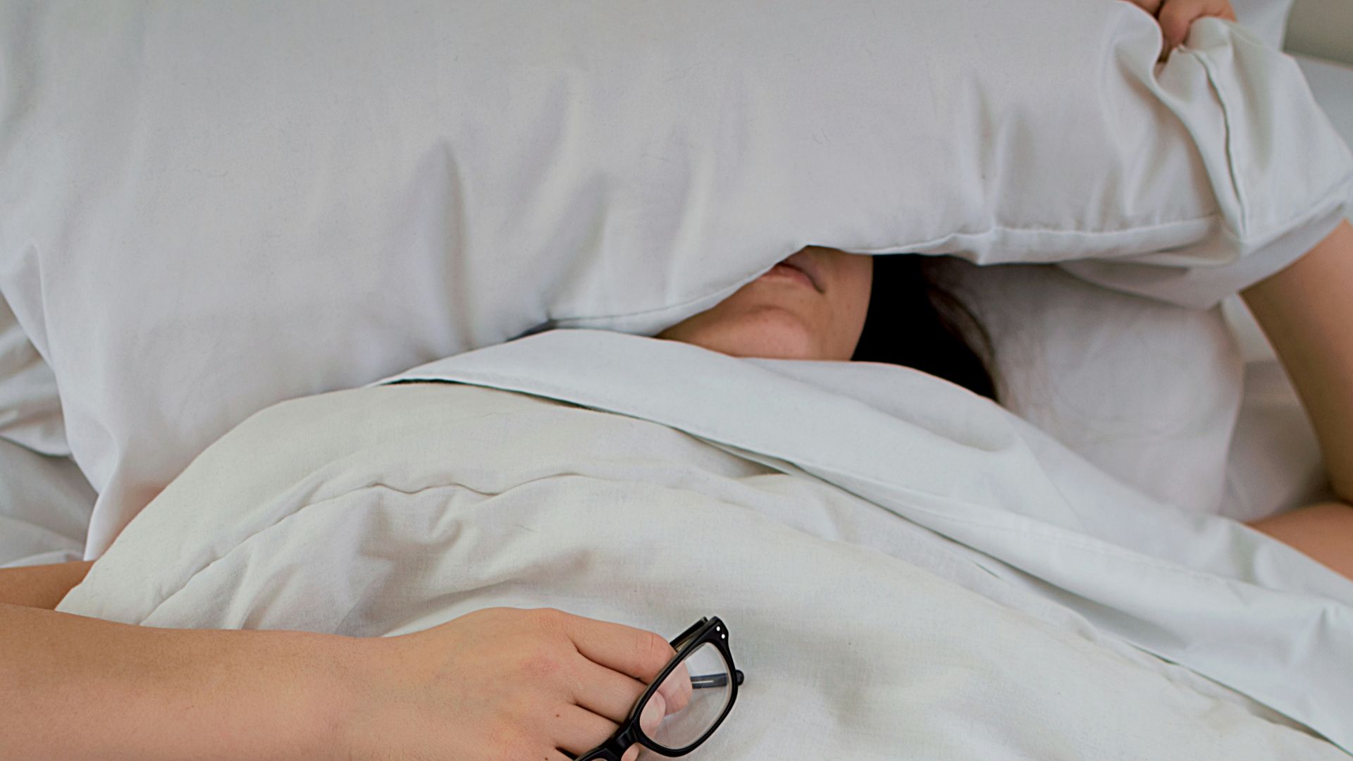 person lying on bed while covering face with pillow and holding eyeglasses
