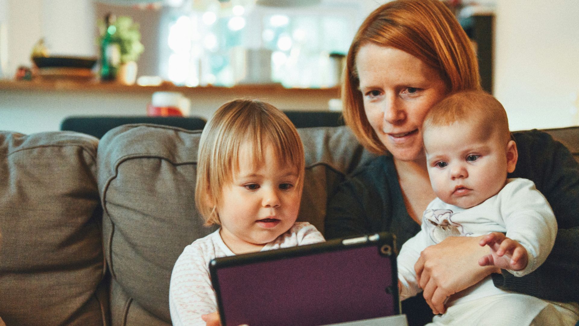 two babies and woman sitting on sofa while holding baby and watching on tablet