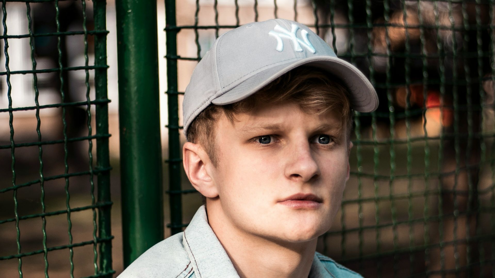 man wearing blue button-up denim jacket sitting beside green fence