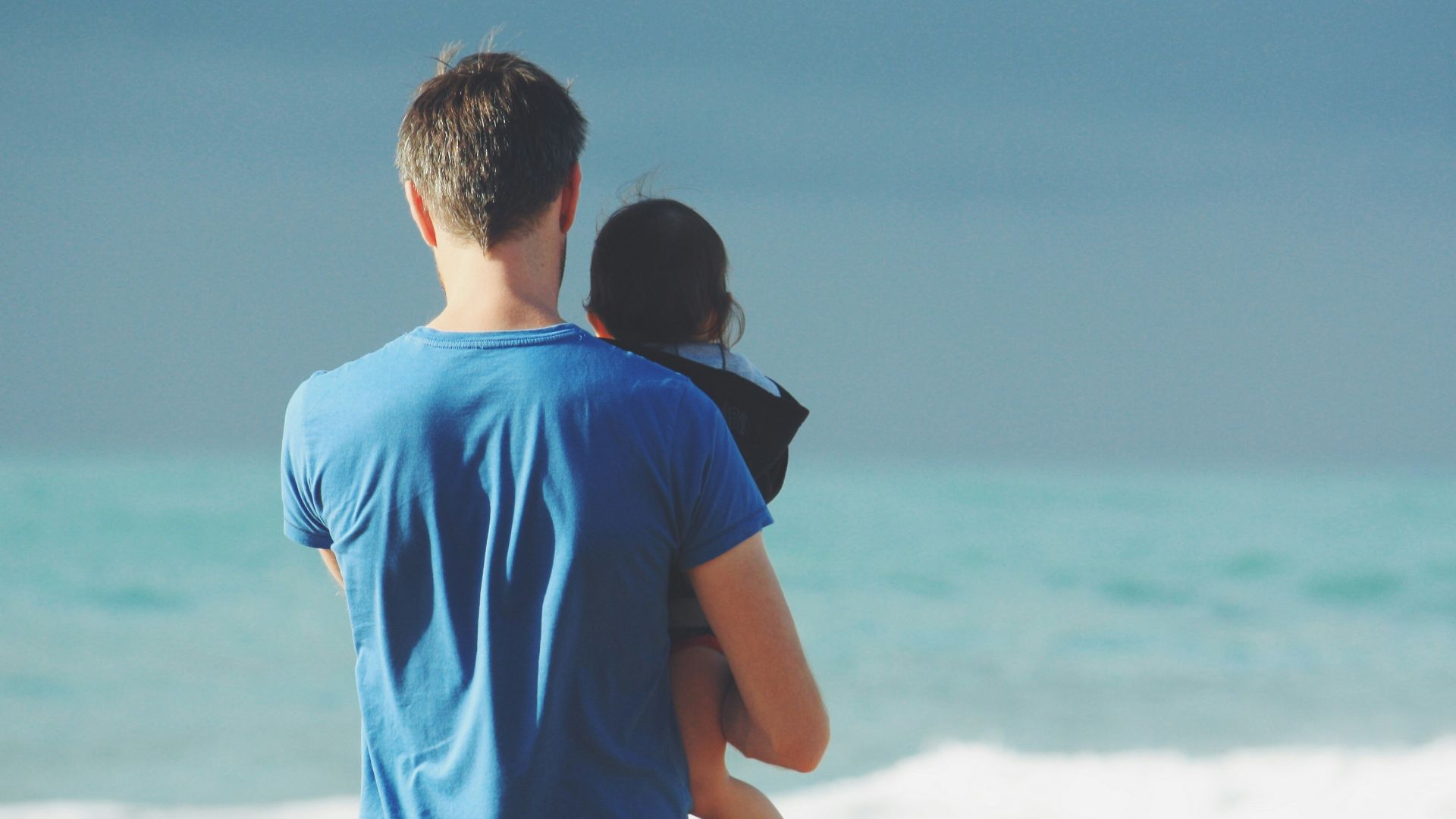 man wears blue crew-neck t-shirt holding toddler wears black hooded jacket near ocean under blue sky at daytime