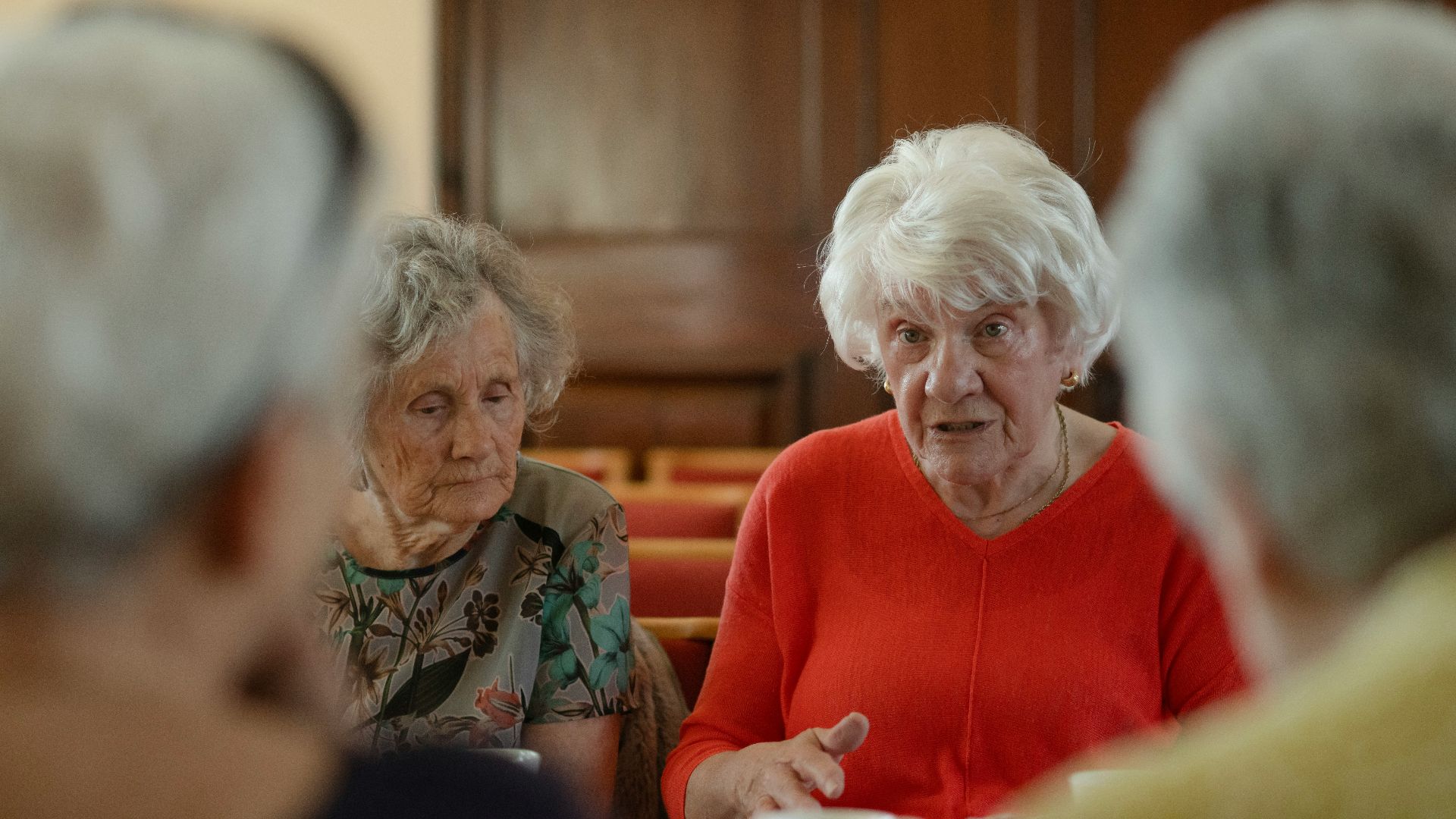 Elderly women sit around a table, talking.