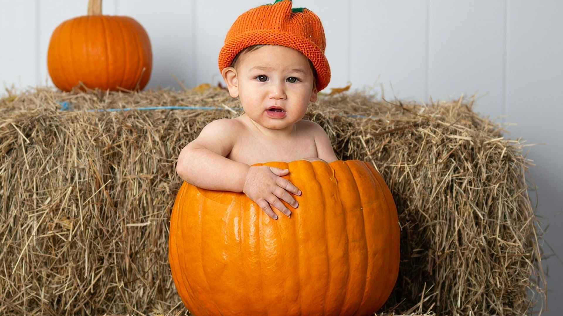 A baby sitting on a hay bale with a pumpkin