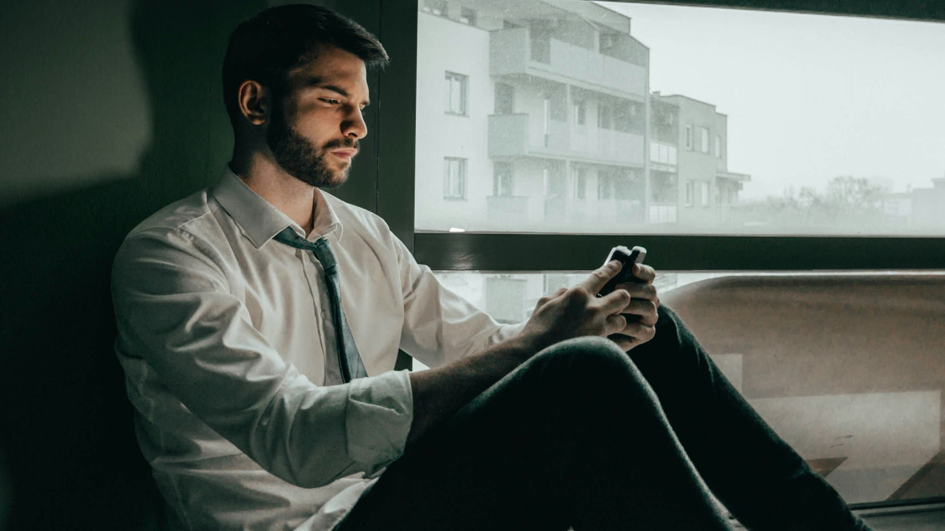 a man sitting on a window sill looking at his cell phone