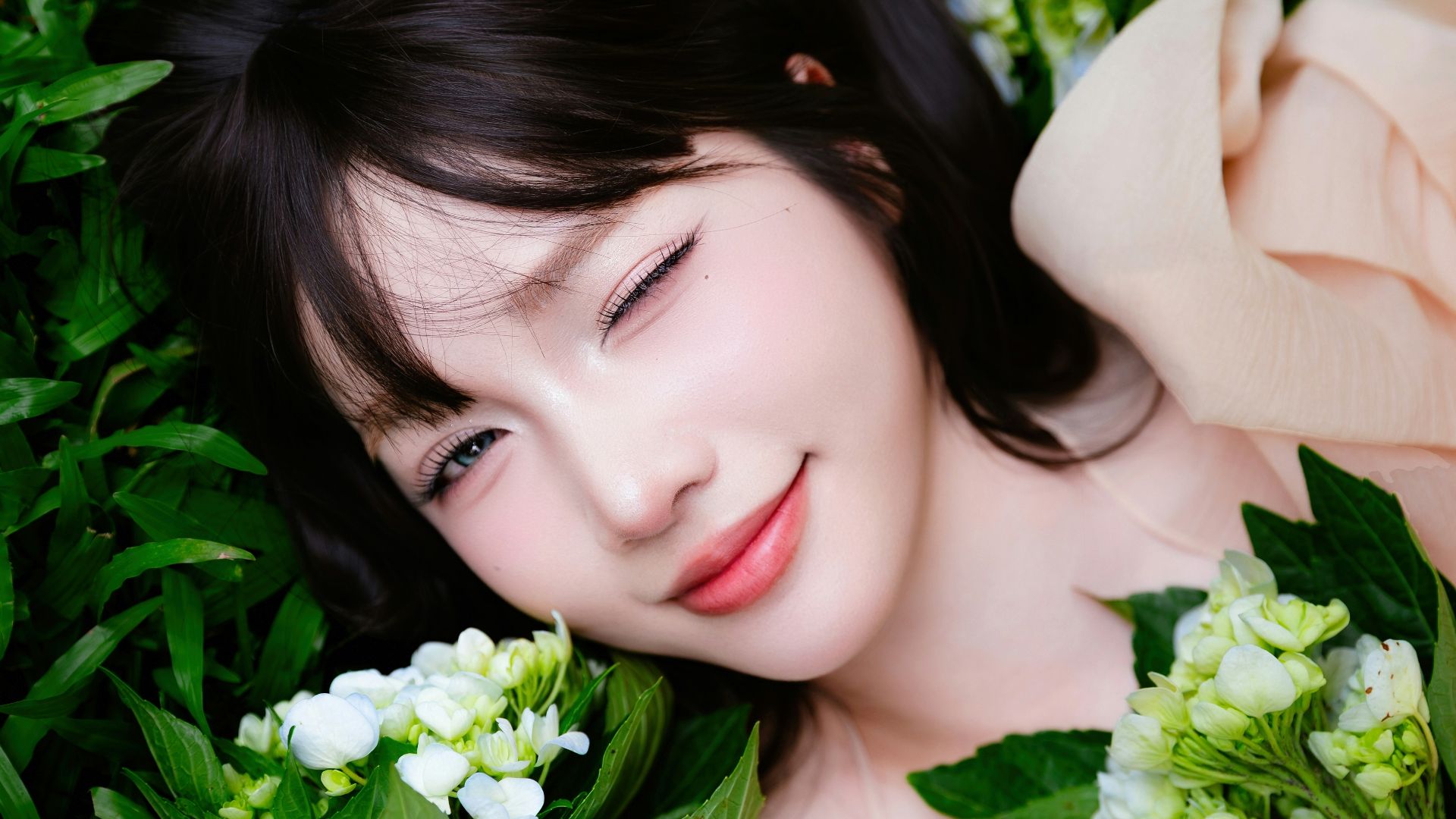 Young woman winks while lying among green leaves and flowers.
