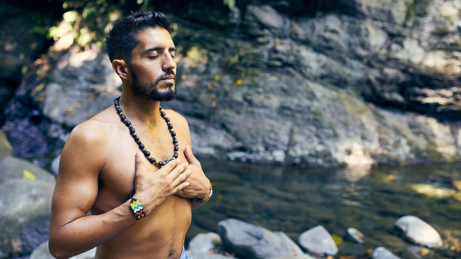 topless man wearing black beaded necklace and blue denim shorts standing on rocky shore during daytime