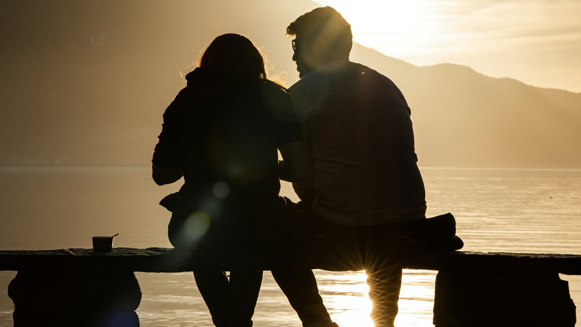 silhouette of man and woman sitting on bench near body of water during sunset