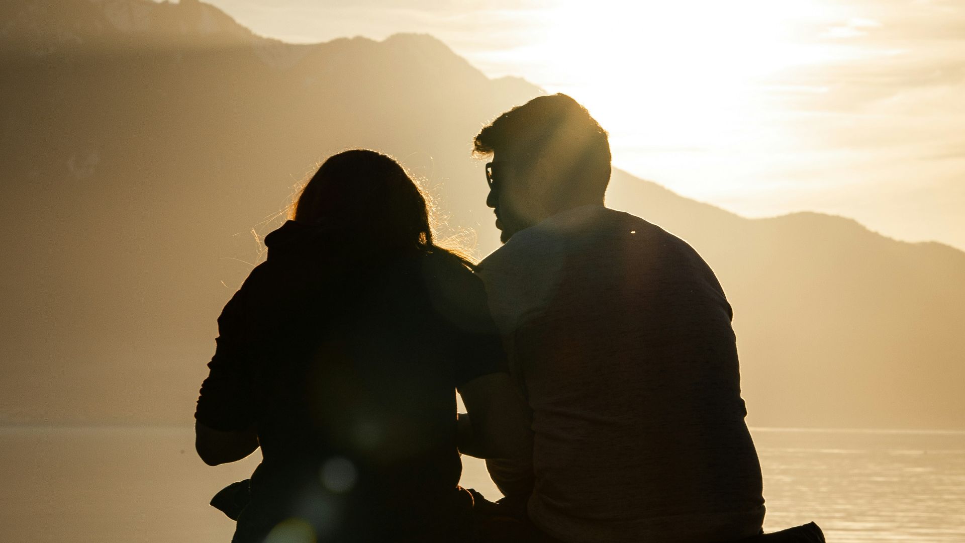 silhouette of man and woman sitting on bench near body of water during sunset