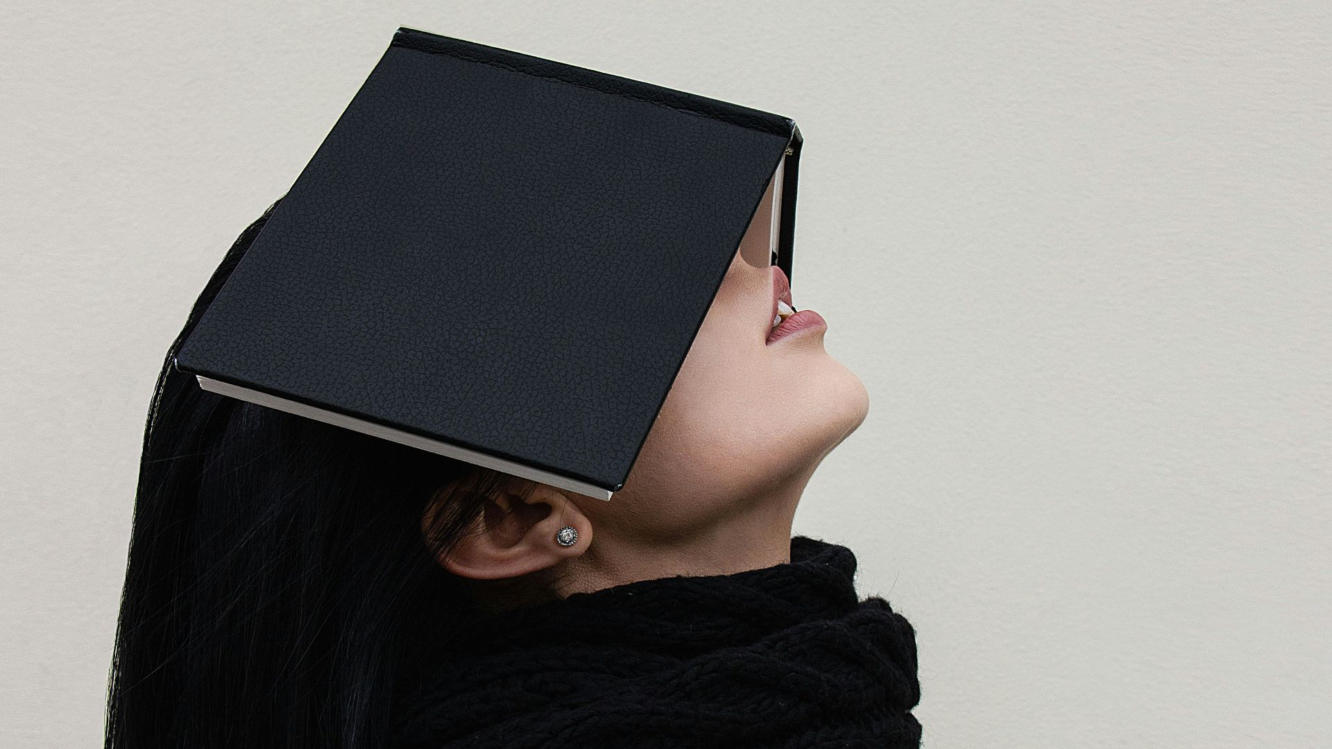 woman in gray sweater and black scarf covering face with black book