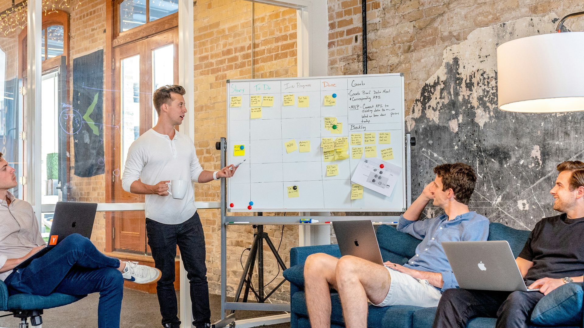 three men sitting while using laptops and watching man beside whiteboard