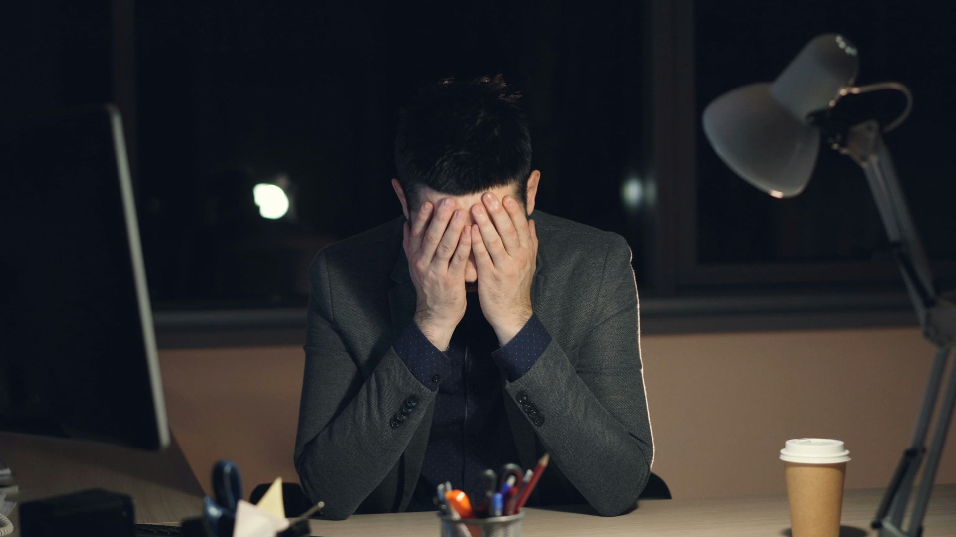 Man in suit sits at desk, head in hands.