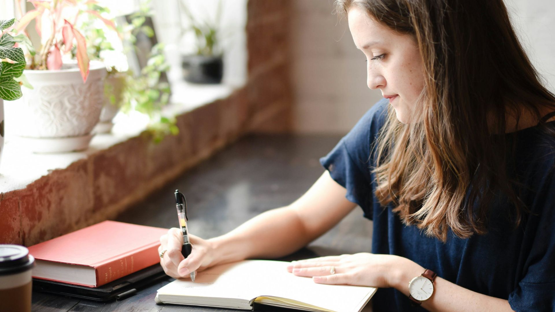 woman sitting in front of black table writing on white book near window