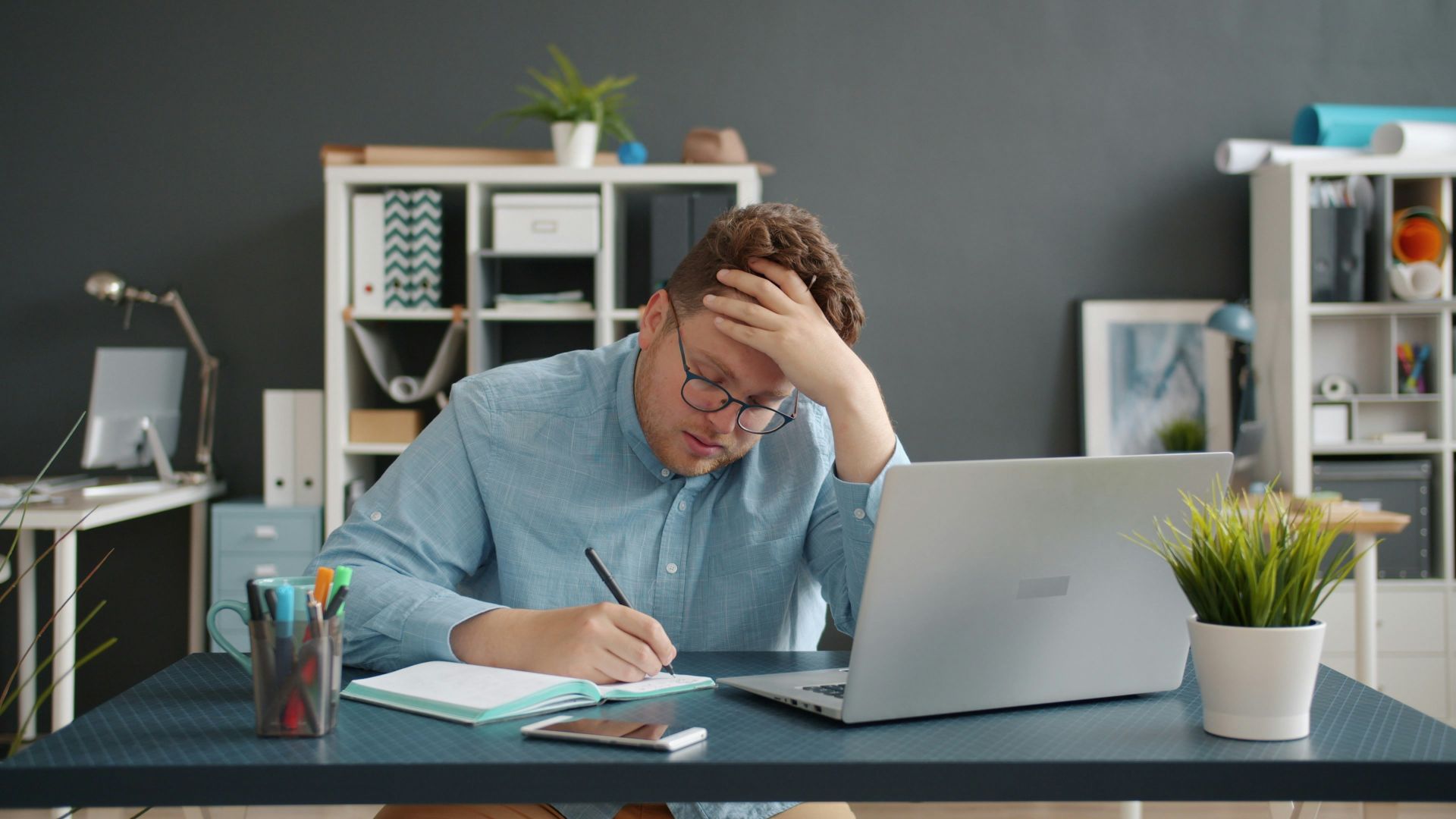 Man writing at desk with laptop, looking stressed.