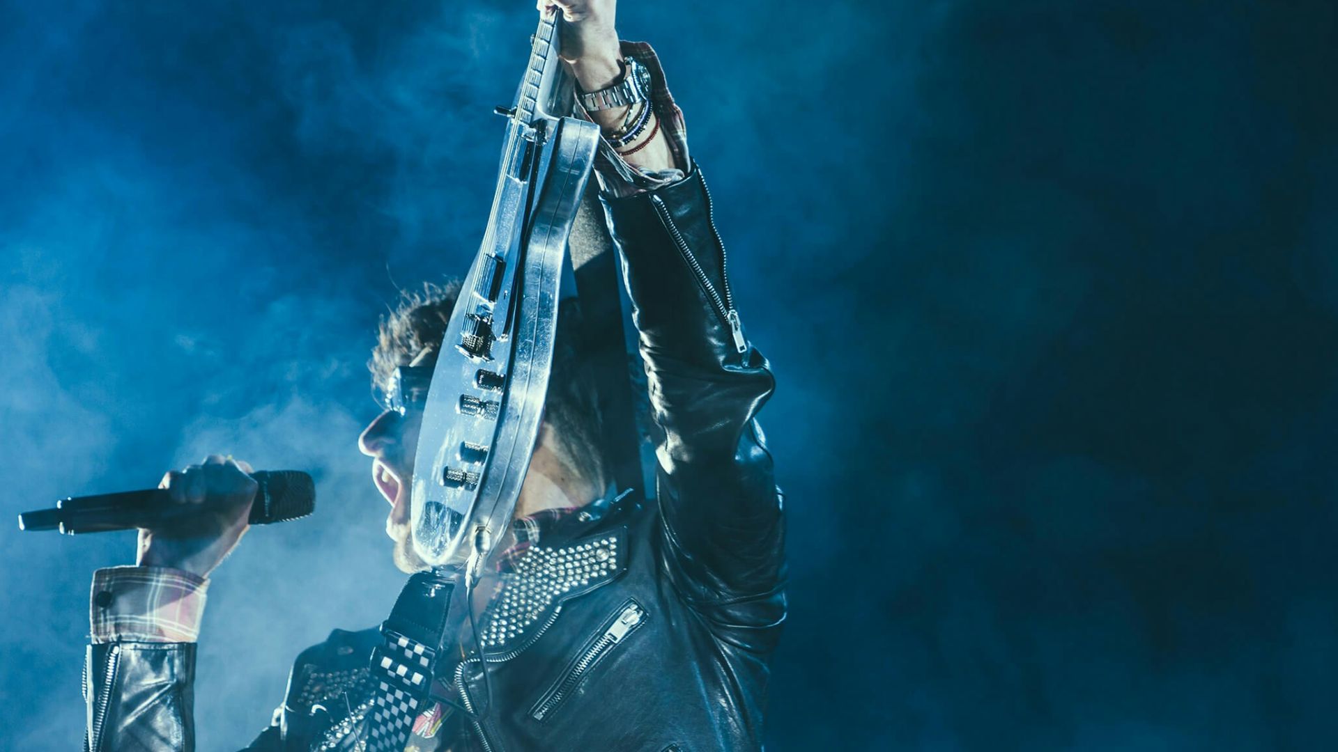 man holding electric guitar and microphone in front of lights inside dim room with smoke
