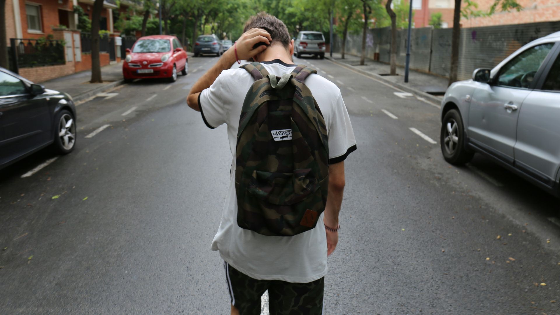 boy wearing white shirt and black shorts carrying backpack standing on black concrete road between vehicles and trees during daytime