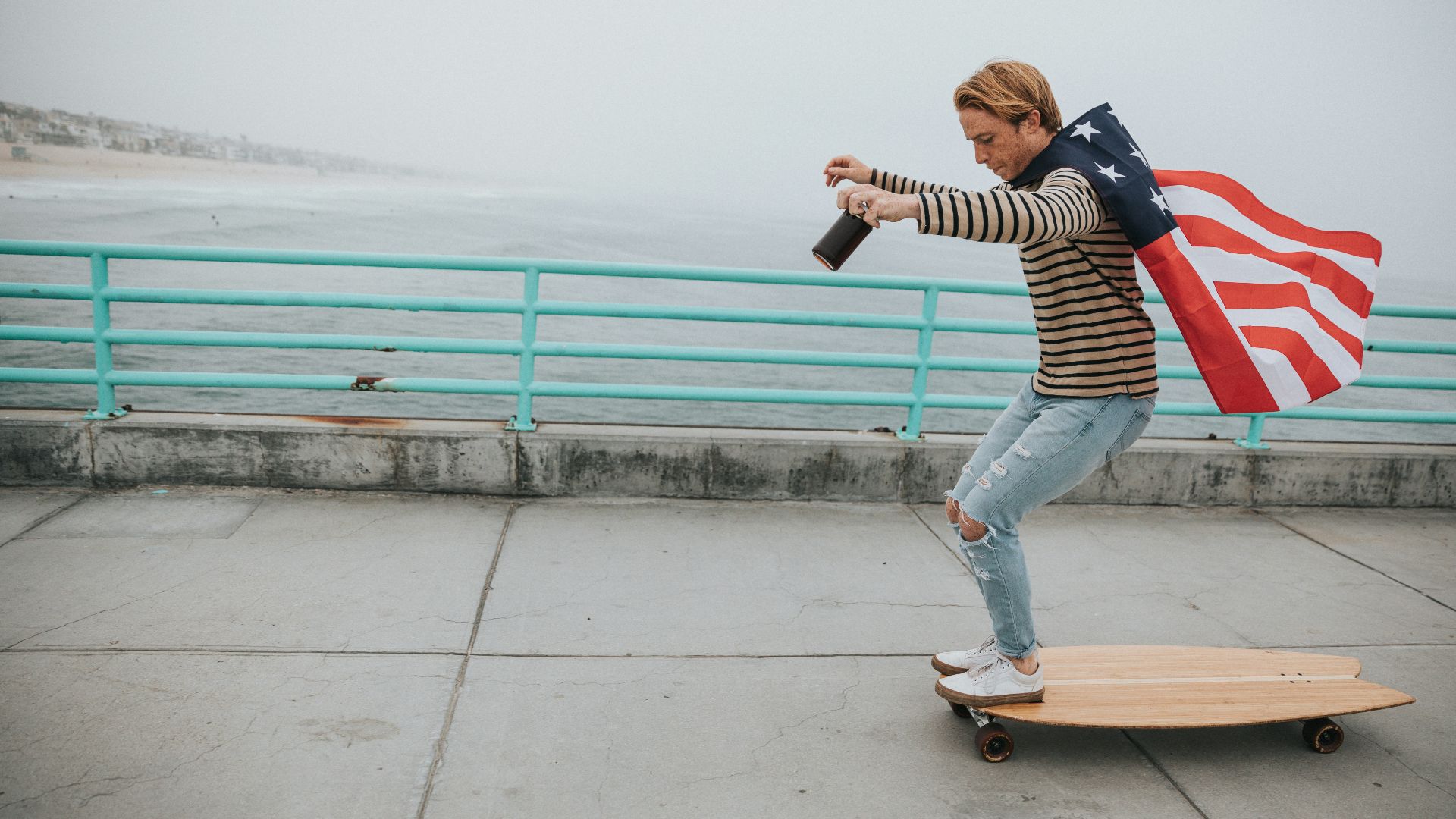 man wearing US flag cape while riding on longboard near body of water during daytime
