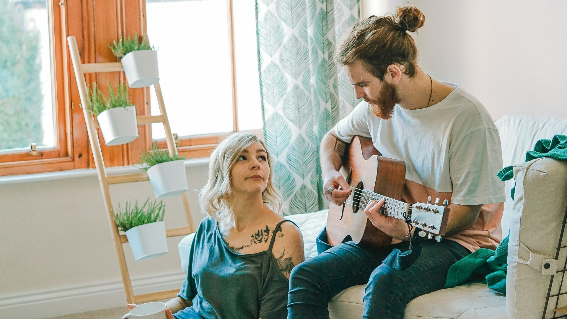 man sitting on sofa playing guitar looking at girl sitting on the ground near window inside room