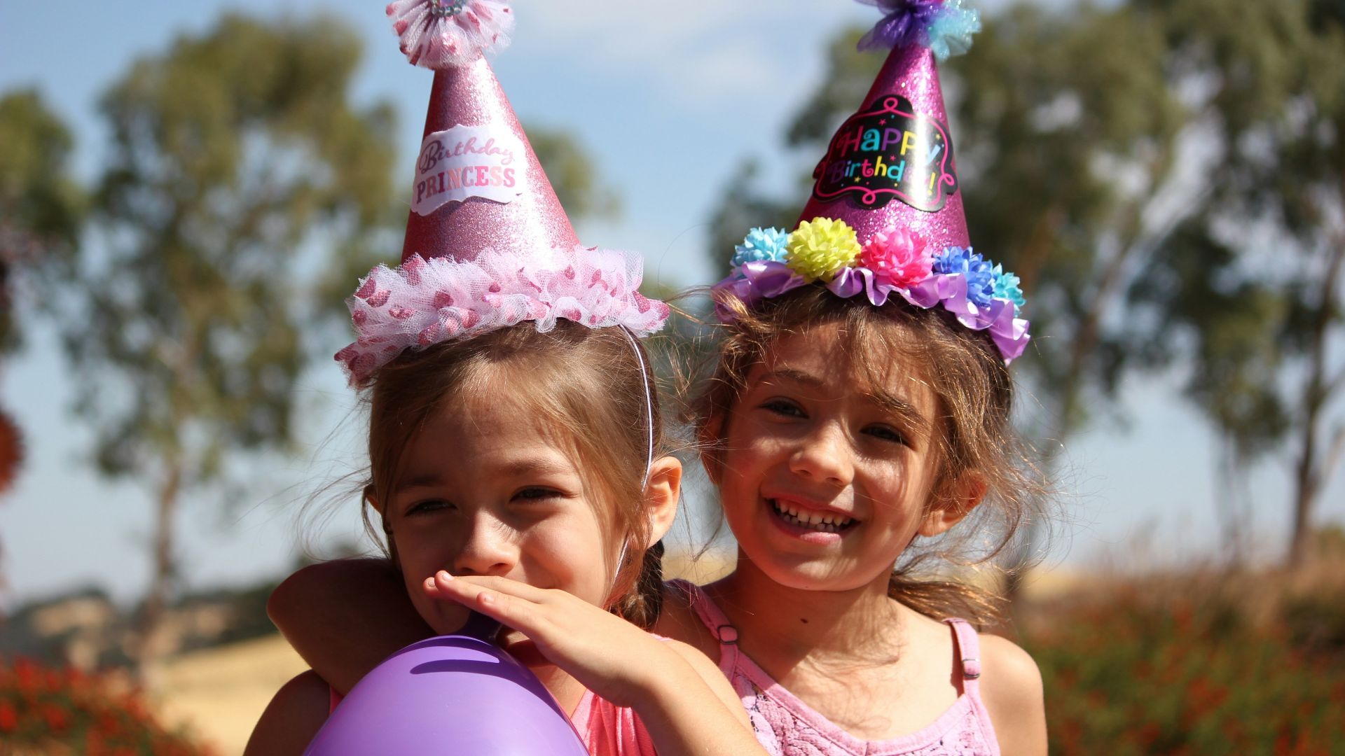 girl in left blowing purple balloon beside girl wearing pink birthday hat