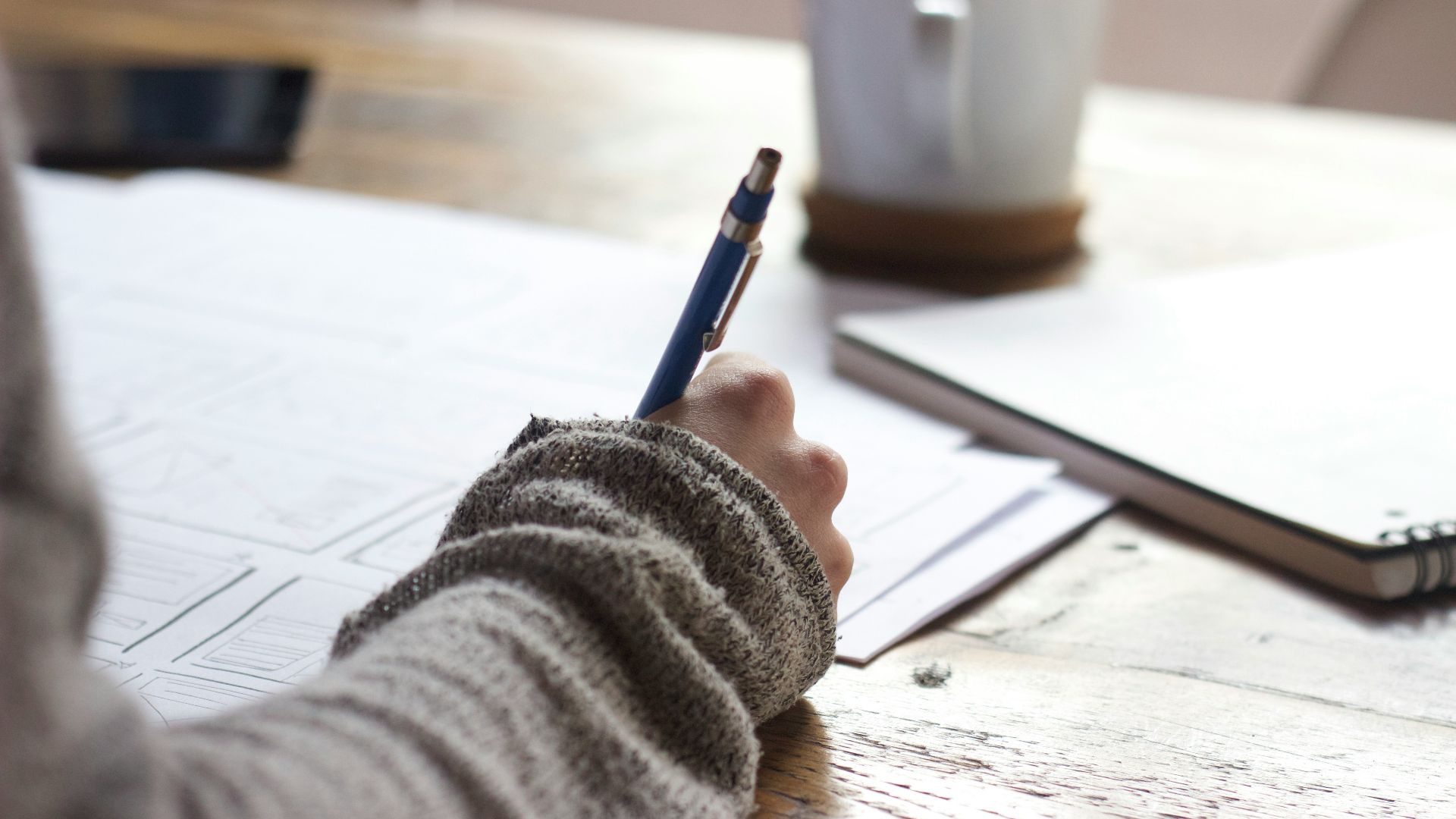 person writing on brown wooden table near white ceramic mug