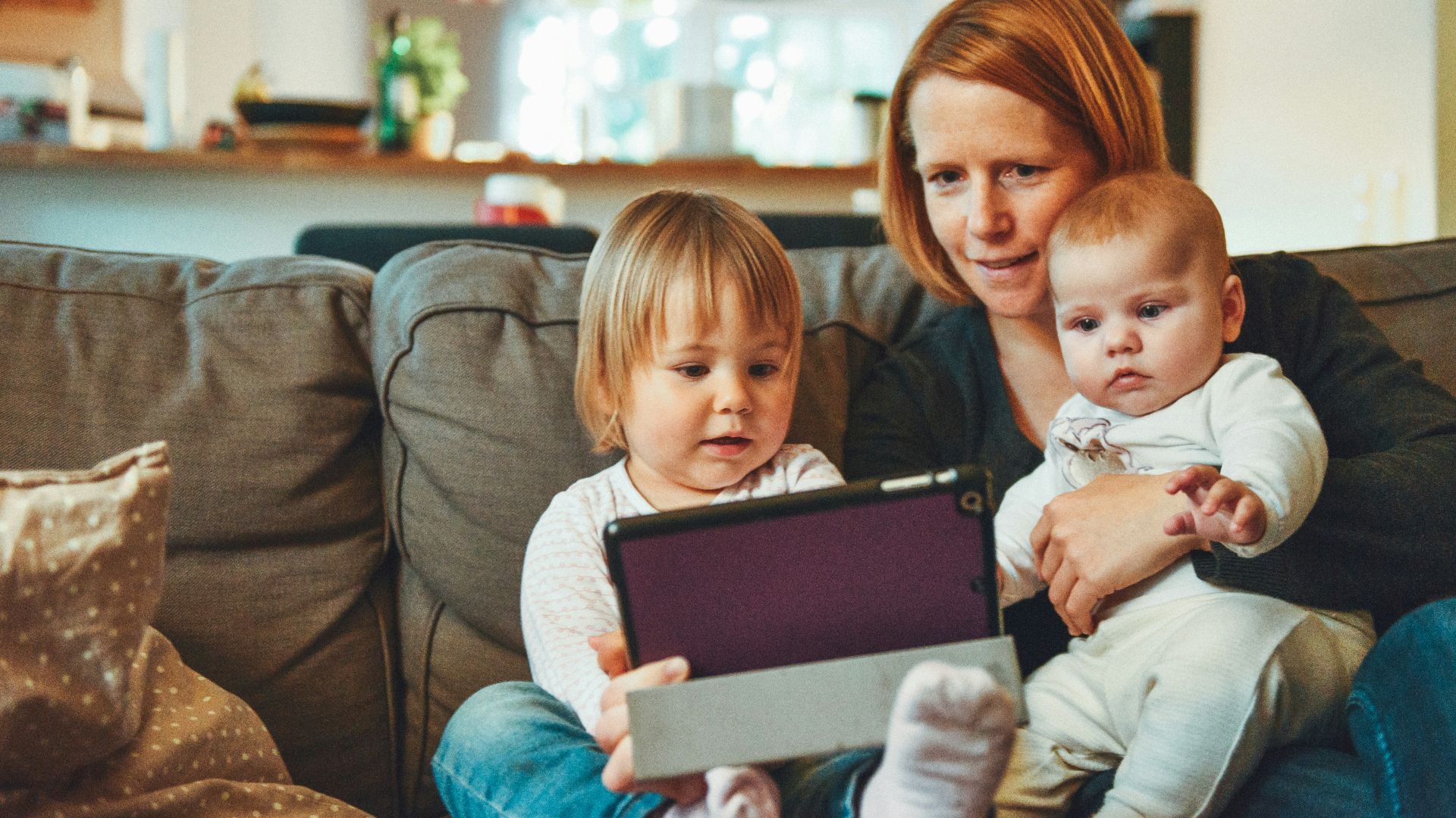 two babies and woman sitting on sofa while holding baby and watching on tablet