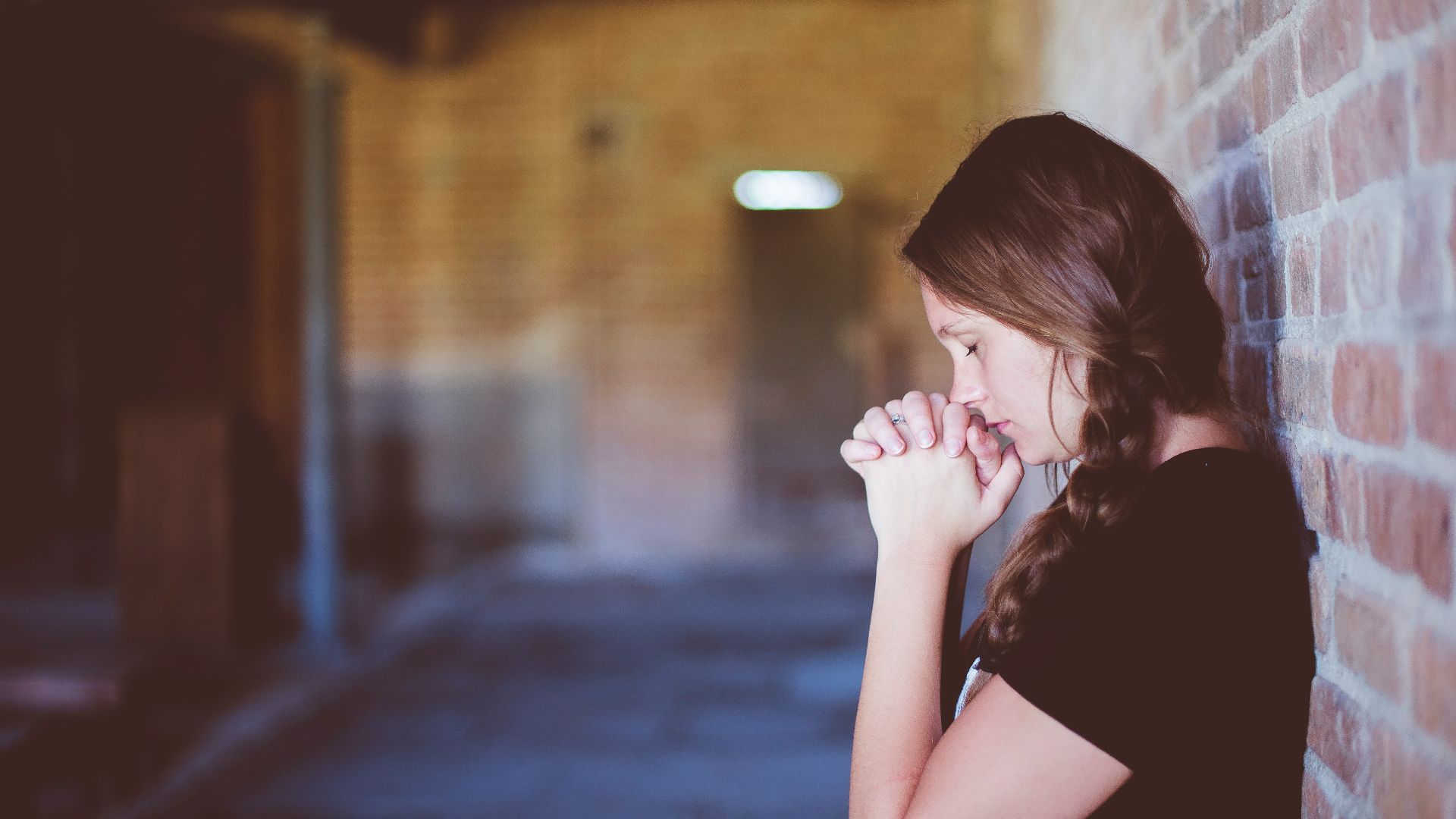 woman praying while leaning against brick wall