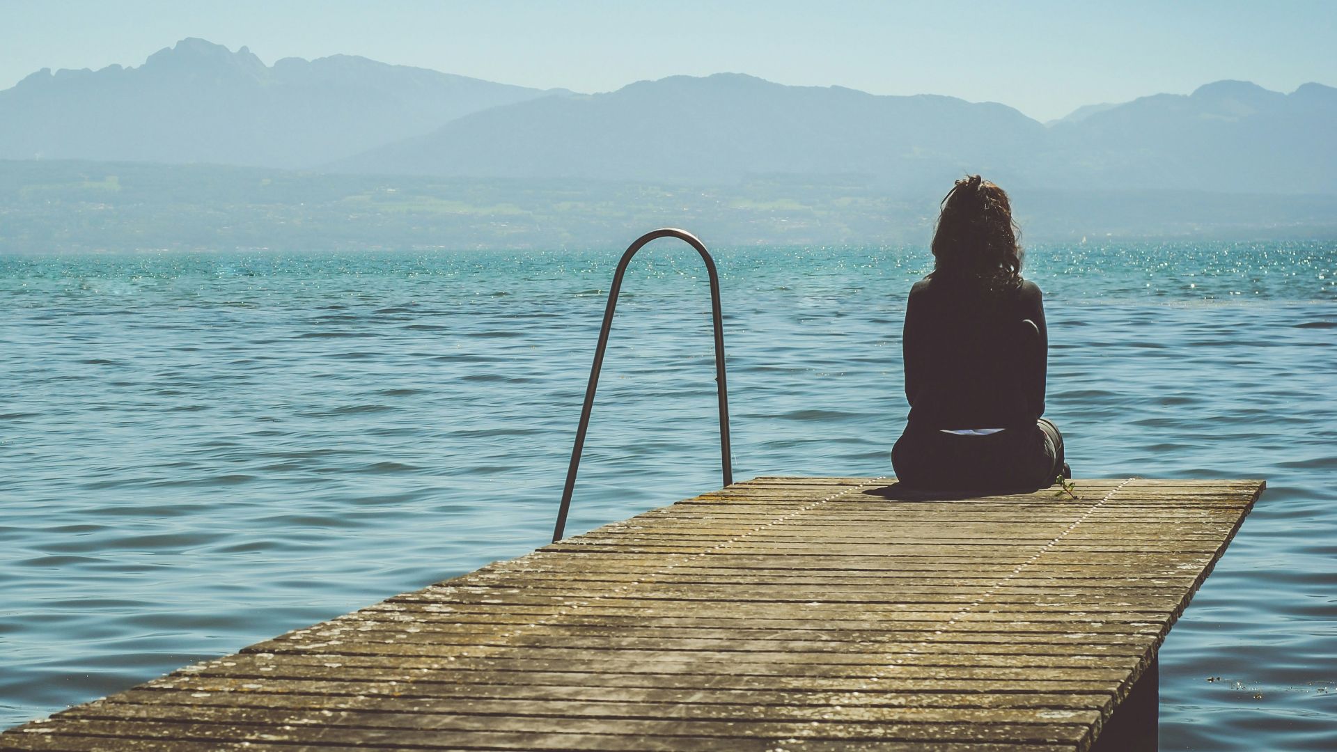 a woman sits on the end of a dock during daytime staring across a lake