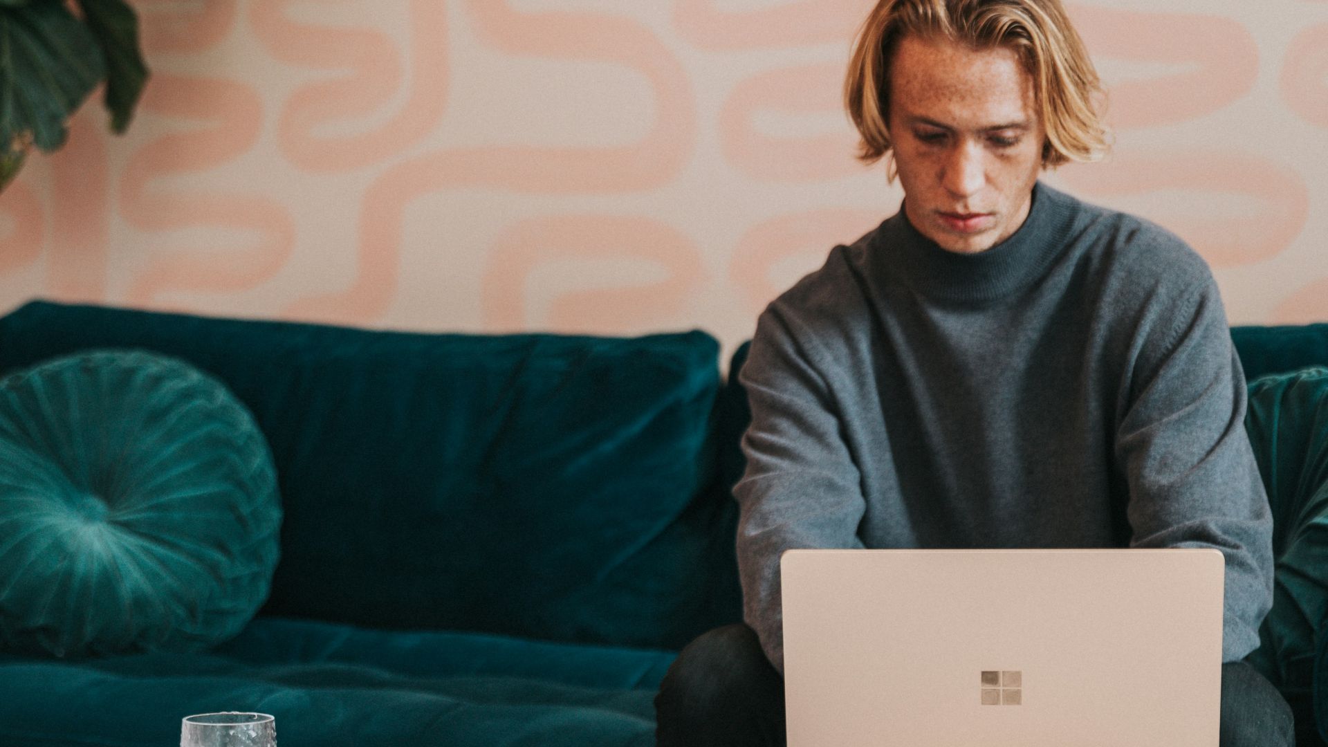 man in gray turtle neck shirt sitting on blue couch using a Microsoft Surface 3 sandstone laptop