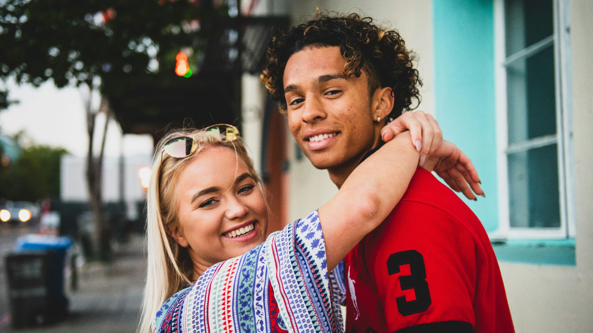 smiling woman with both hands wrap around man's neck near white concrete building