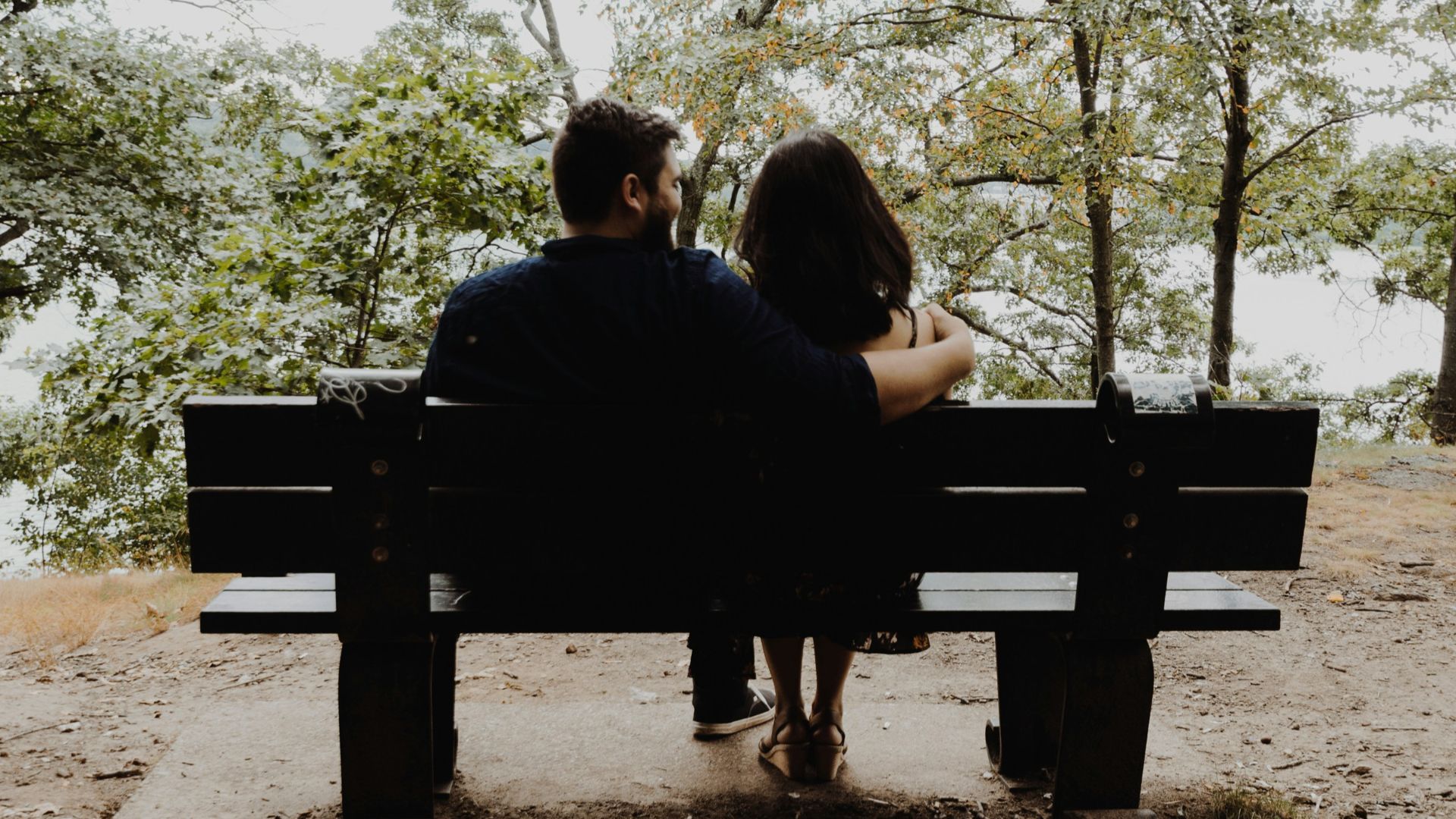 man looking to woman sitting on black wooden bench in front of tall trees during daytime