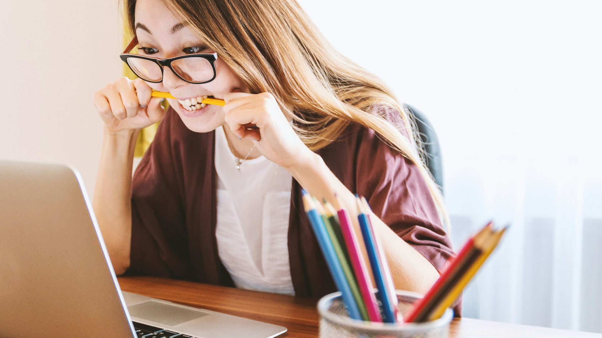 woman biting pencil while sitting on chair in front of computer during daytime
