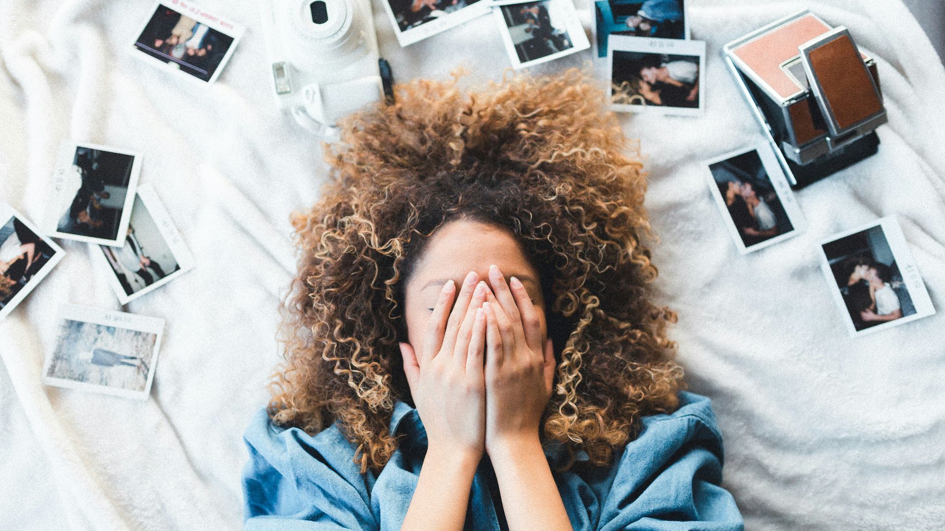 woman lying on bed covering her face surrounded by photos and white camera