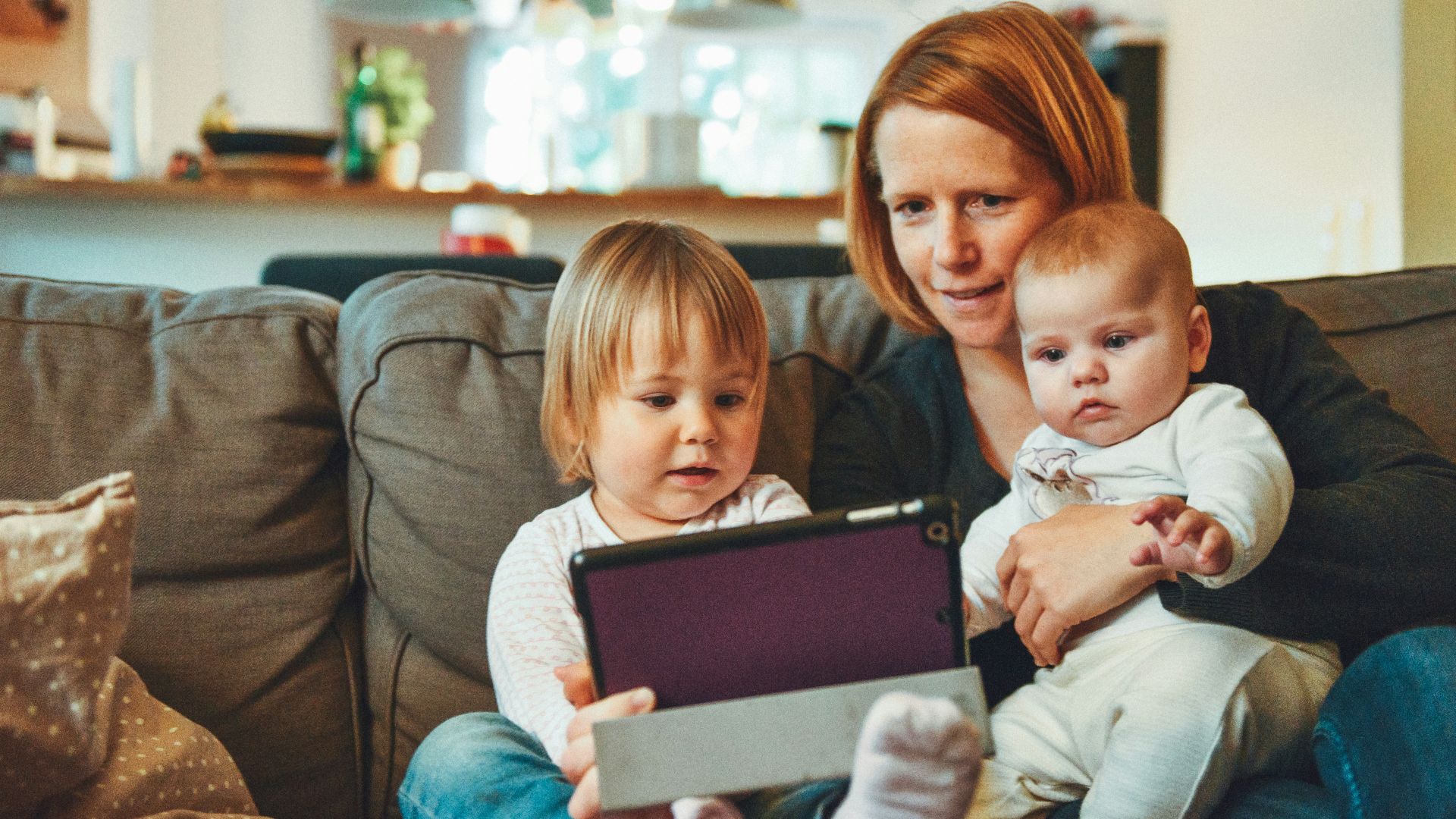 two babies and woman sitting on sofa while holding baby and watching on tablet