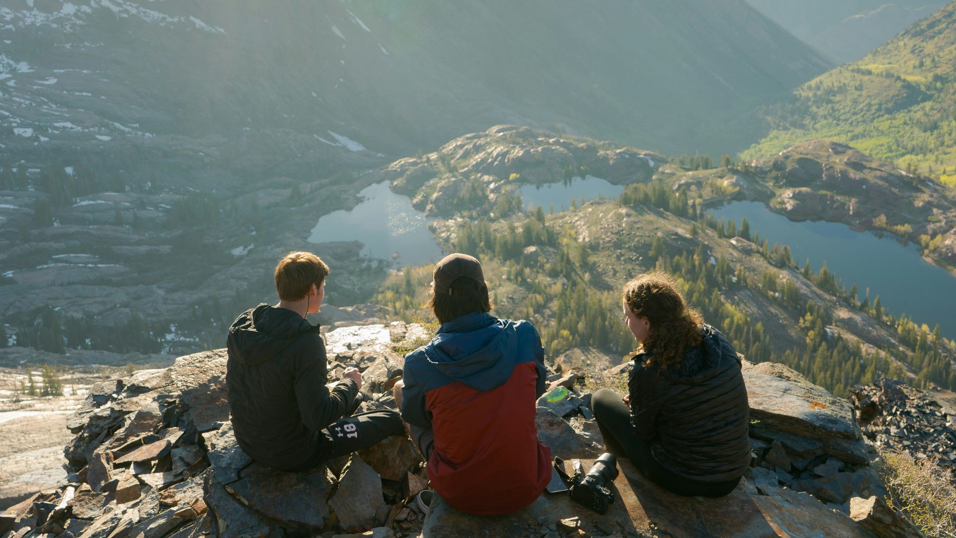 two men and one woman sitting on cliff overlooking lake and mountains during daytime